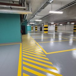 EPOXYPLANET Posadzki żywiczne Kamil Burda - Szara posadzka w garażu podziemnym z żółtymi pasami i oznaczeniami. Widoczne rury pod sufitem i słupy w czarno-żółte pasy. Ściana w kolorze morskim.