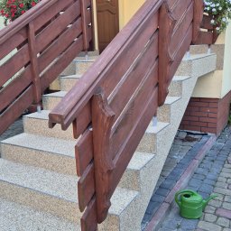 EPOXYPLANET Posadzki żywiczne Kamil Burda - Schody wejściowe z drewnianą balustradą, wykończone jasnym, drobnym kamieniem. Na dole zielona konewka, w tle kwiaty w oknach i drzwi wejściowe.