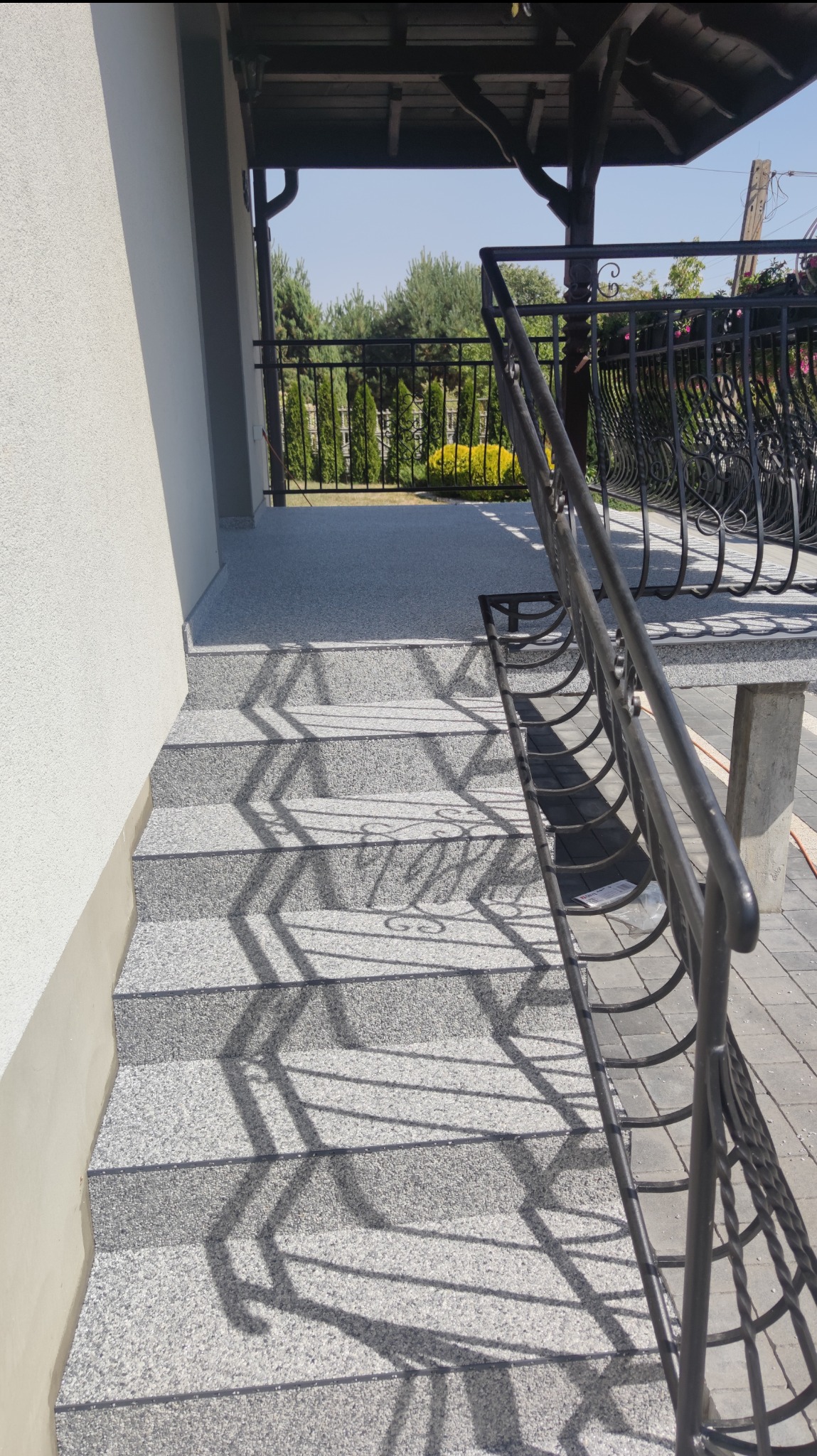 Schody zewnętrzne pokryte szarą posadzką żywiczną typu kamienny dywan, z czarną metalową balustradą, w słoneczny dzień.