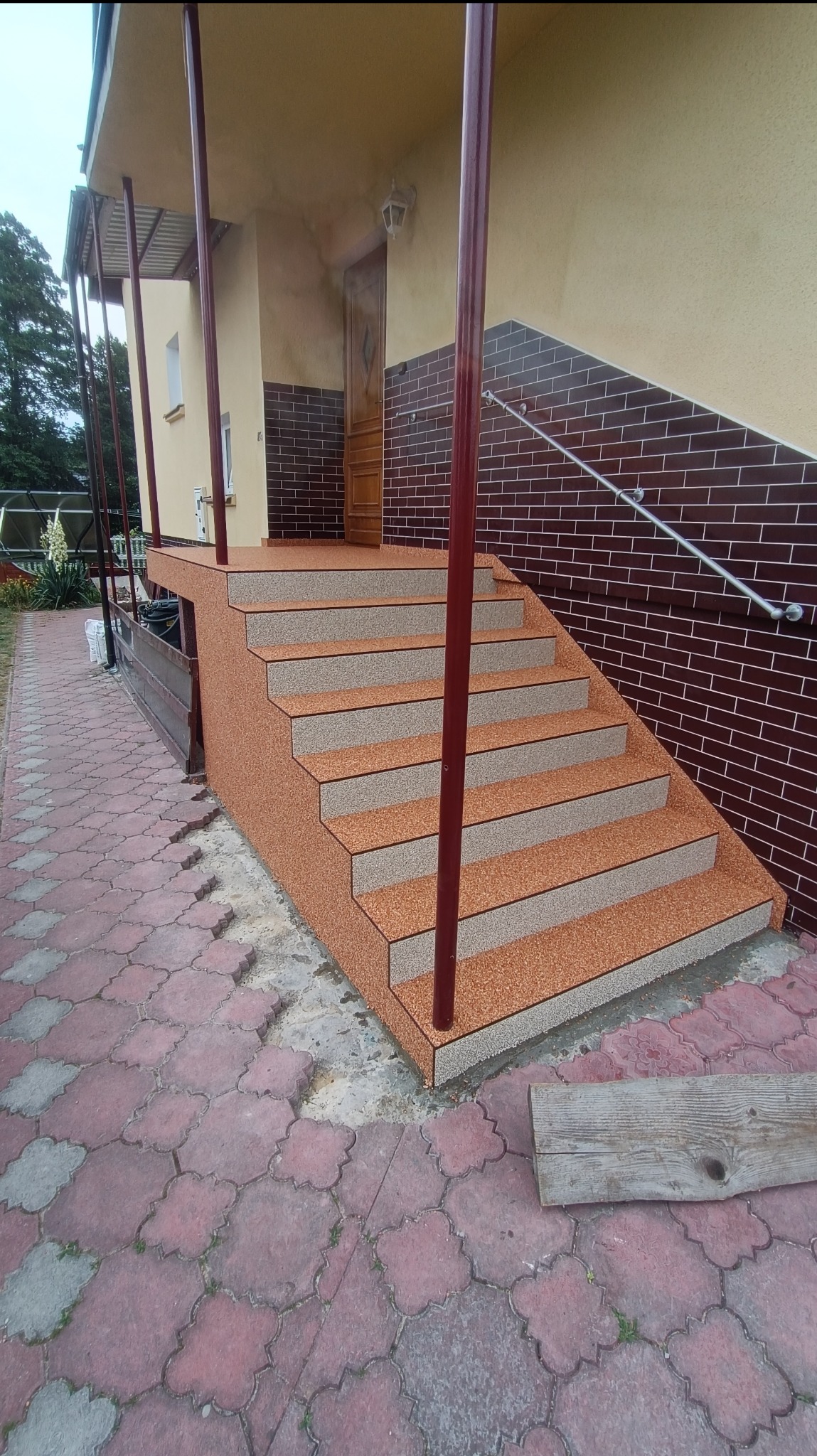 Schody wejściowe do domu z kamiennym dywanem w odcieniach brązu i beżu, wykończone listwami. Widoczna ceglana elewacja budynku i fragment chodnika z kostki brukowej.