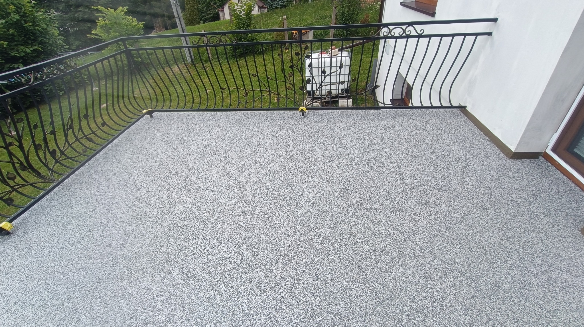 Szary kamienny dywan na balkonie z kutą, czarną balustradą z motywem roślinnym, widok na trawnik i budynek w tle.