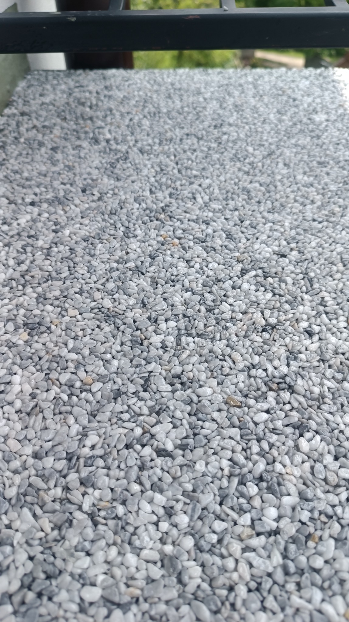 Szara powierzchnia wykonana z drobnych kamyczków, tworząca efekt kamiennego dywanu na balkonie, widoczna z bliska, z fragmentem metalowej barierki w górnej części kadru.
