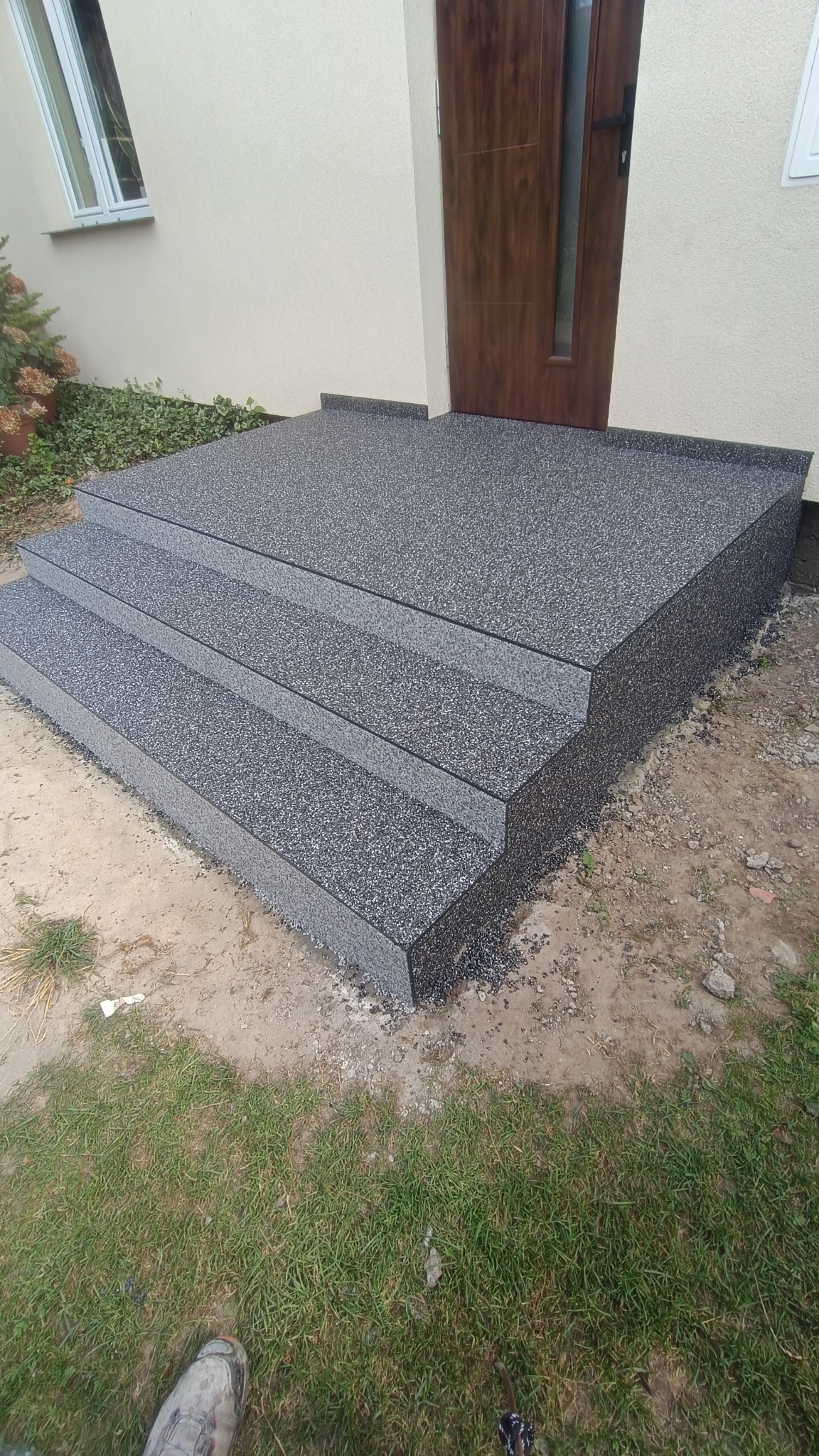 Zewnętrzne schody wejściowe wykończone szarą, kamienną posadzką żywiczną, prowadzące do brązowych drzwi wejściowych.