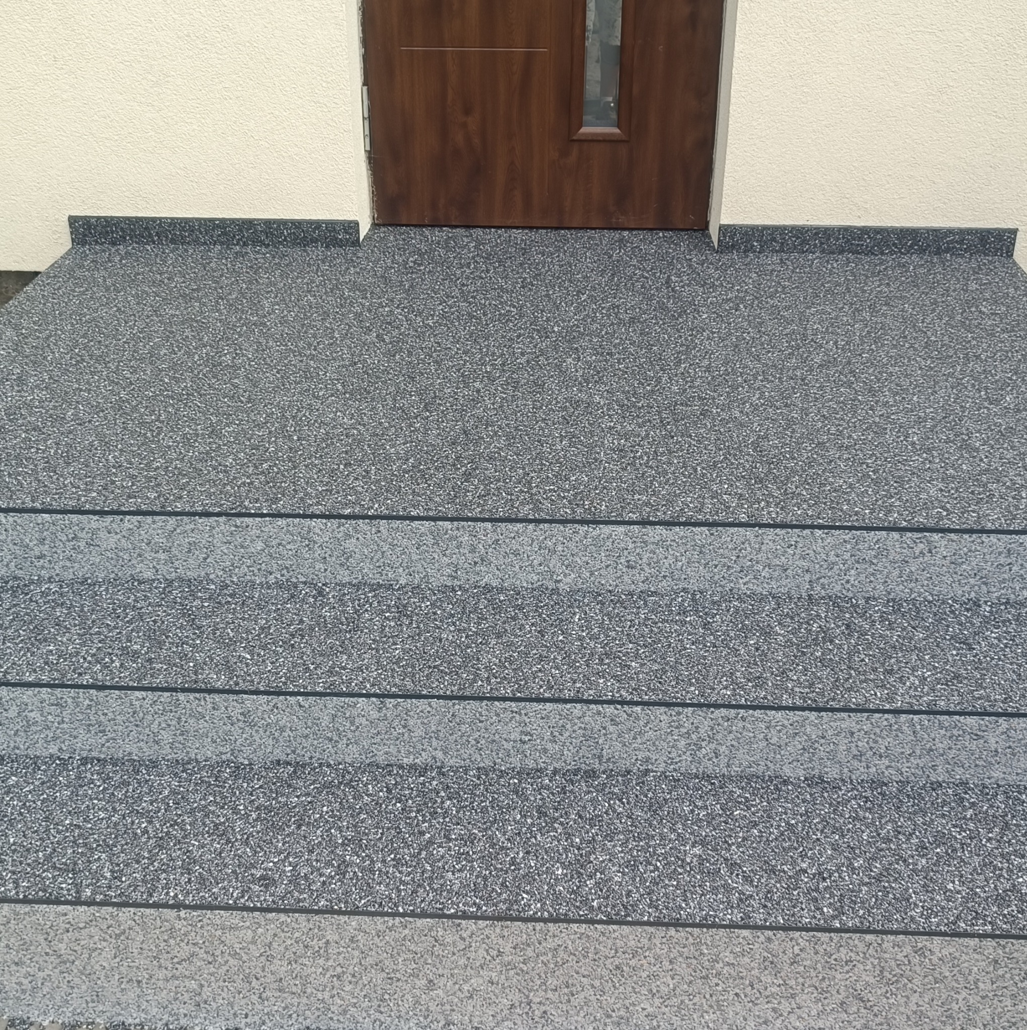 Schody wejściowe wykończone szarą, kamienną posadzką żywiczną typu kamienny dywan, prowadzące do brązowych drzwi wejściowych.