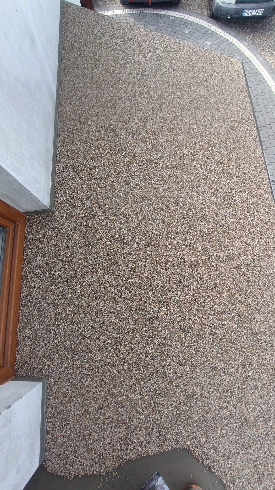 Beżowa posadzka z kamiennego dywanu przylegająca do białej ściany budynku i kostki brukowej pod zaparkowanymi samochodami.