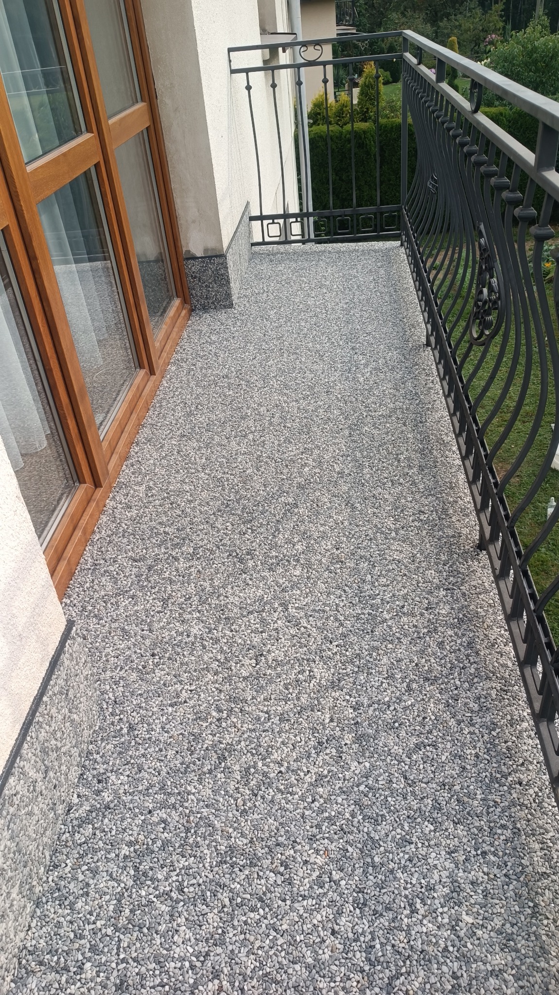 Balkon z szarą posadzką kamienny dywan, widok na zewnątrz przez ozdobną metalową balustradę, w tle zieleń.