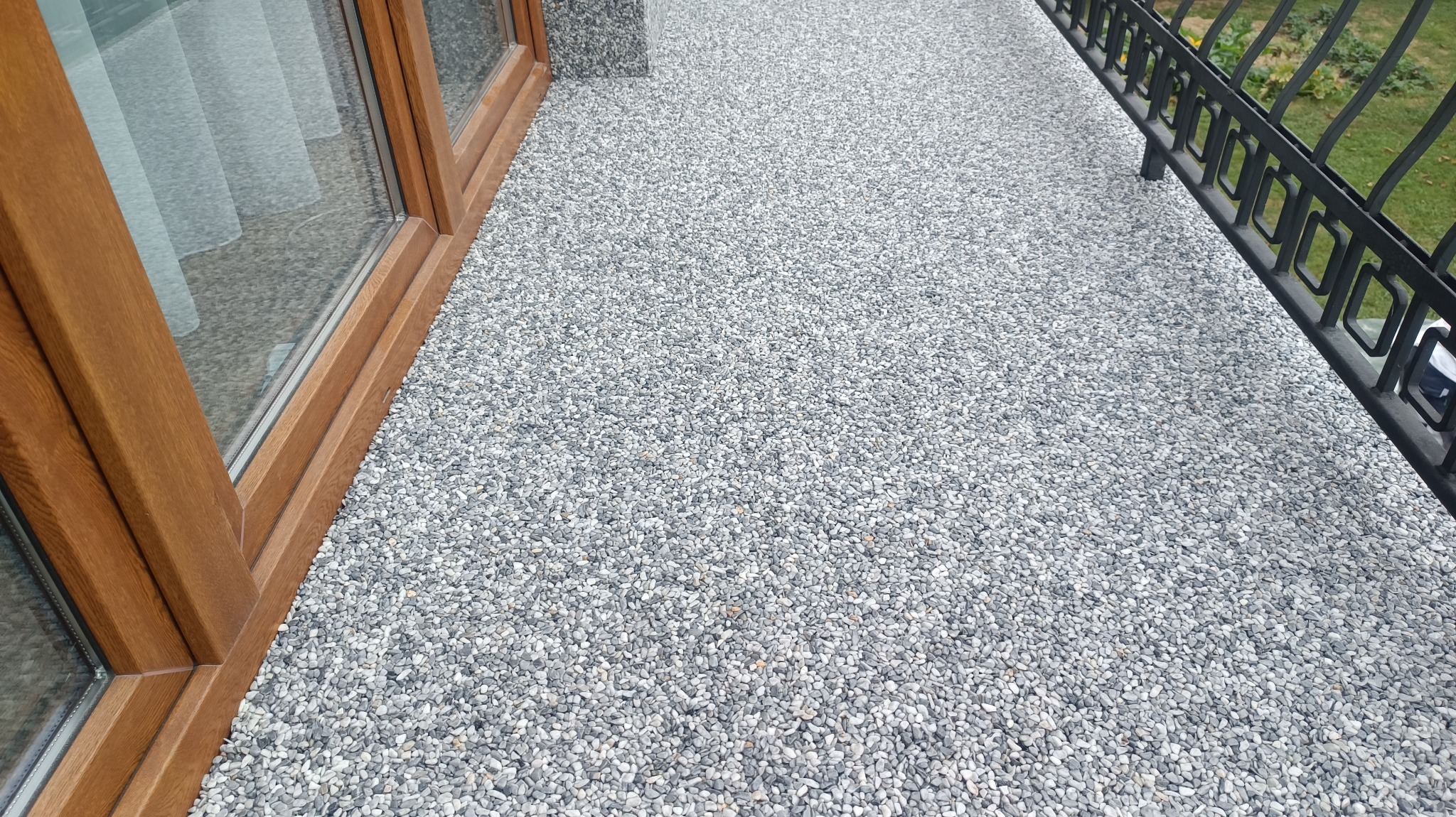 Szary kamienny dywan na balkonie, widoczna drewniana rama okienna i czarna metalowa balustrada