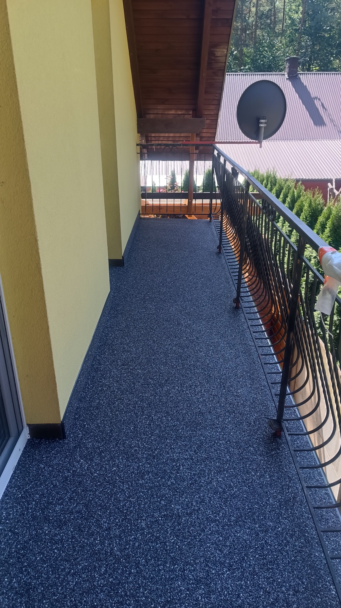 Balkon z posadzką żywiczną typu kamienny dywan w odcieniach szarości i błękitu, z widokiem na zieleń drzew i dach sąsiedniego budynku z anteną satelitarną.