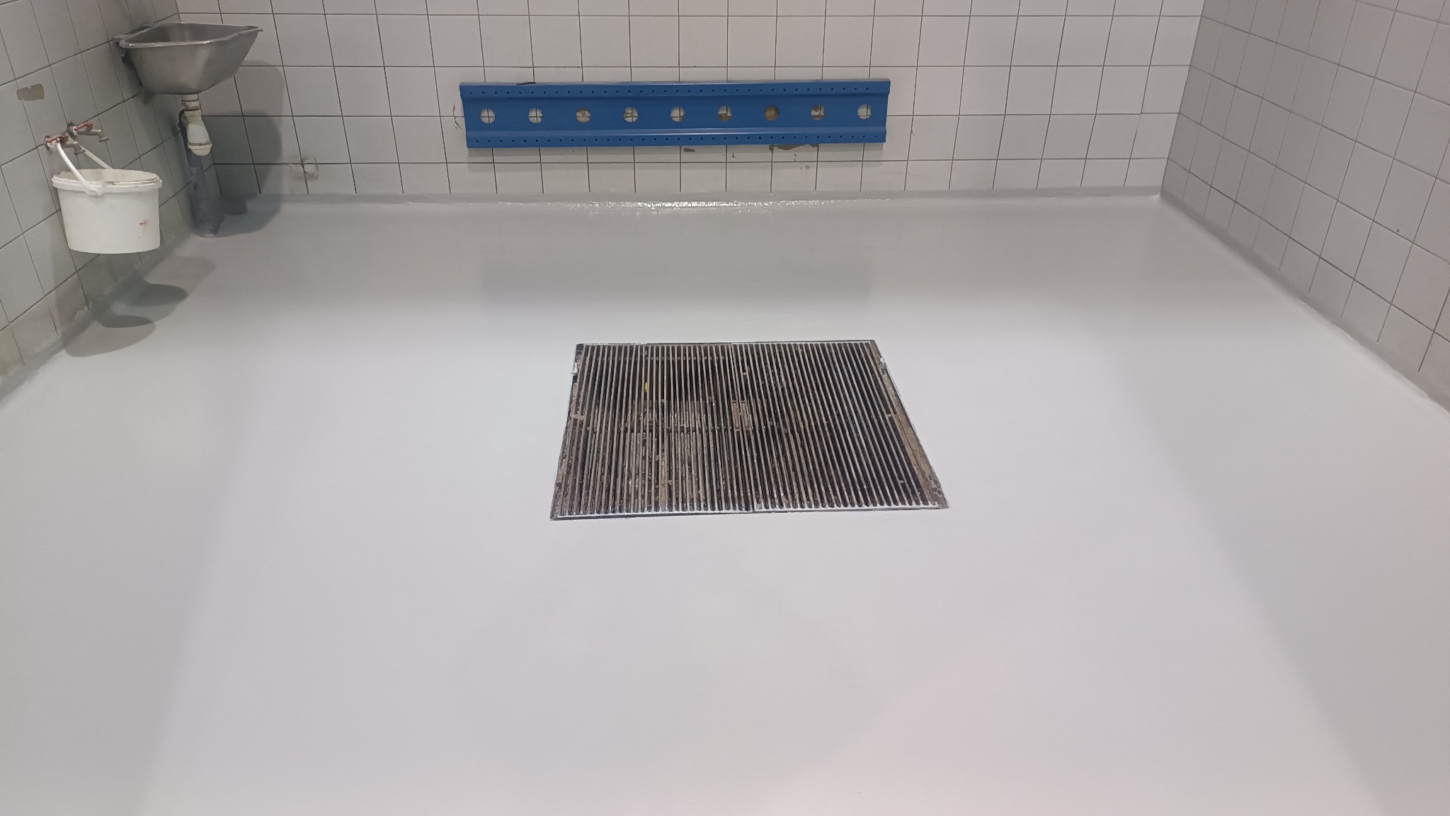 Świeżo wylana, jasnoszara posadzka żywiczna w pomieszczeniu z białymi płytkami ściennymi i odpływem liniowym; widoczny także biały pojemnik z narzędziami oraz niebieski element mocujący na ścianie.