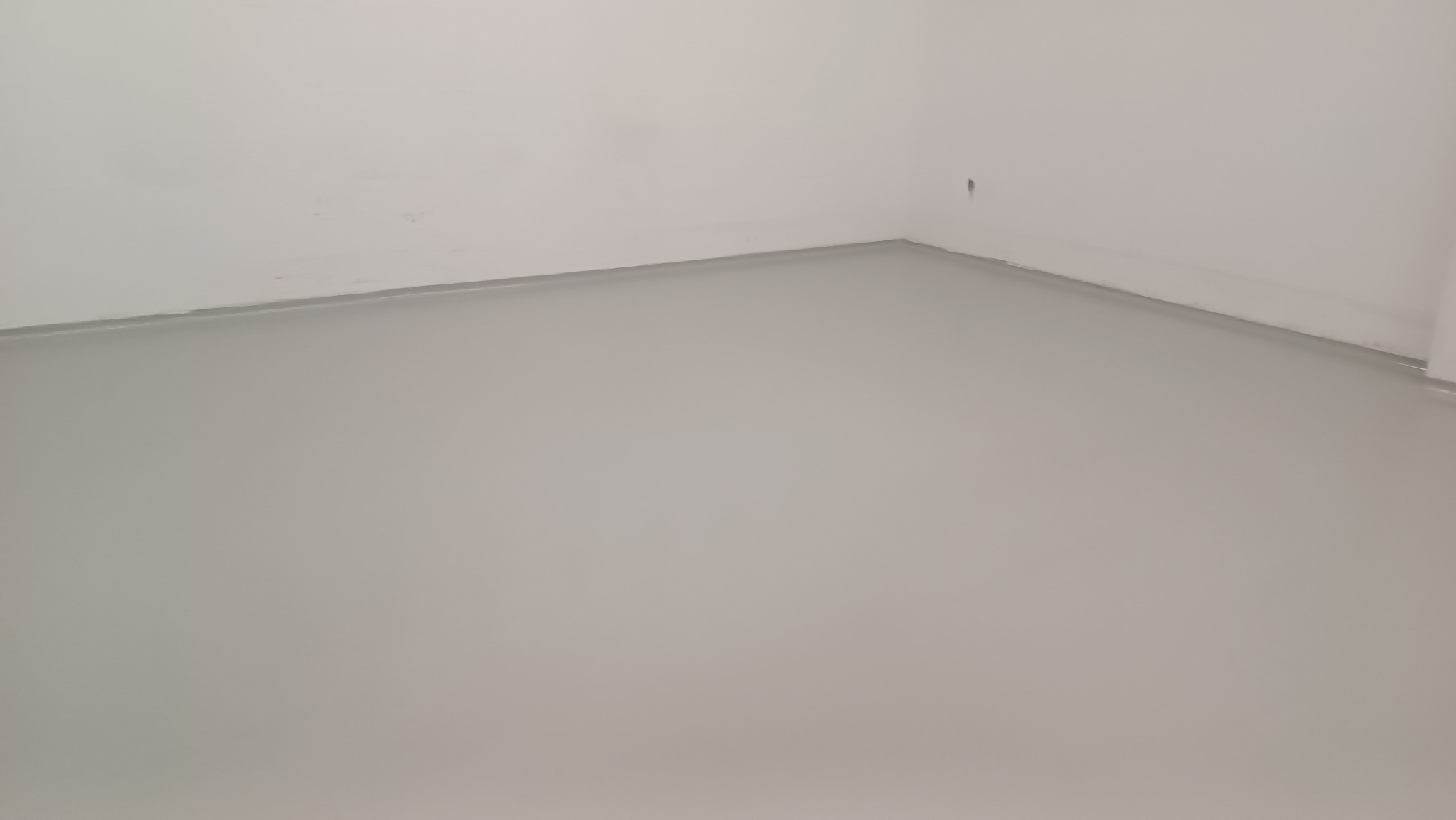 Gładka, szara posadzka w narożniku pomieszczenia z białymi ścianami. Widoczna subtelna listwa przypodłogowa.