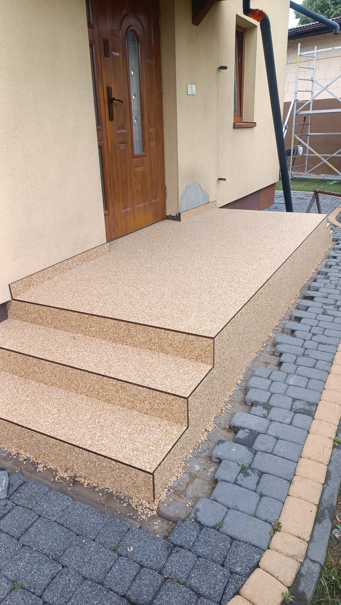 Wykończone schody wejściowe do domu pokryte beżową, drobną posadzką kamienną, z widocznymi detalami wykończenia i kostką brukową obok.