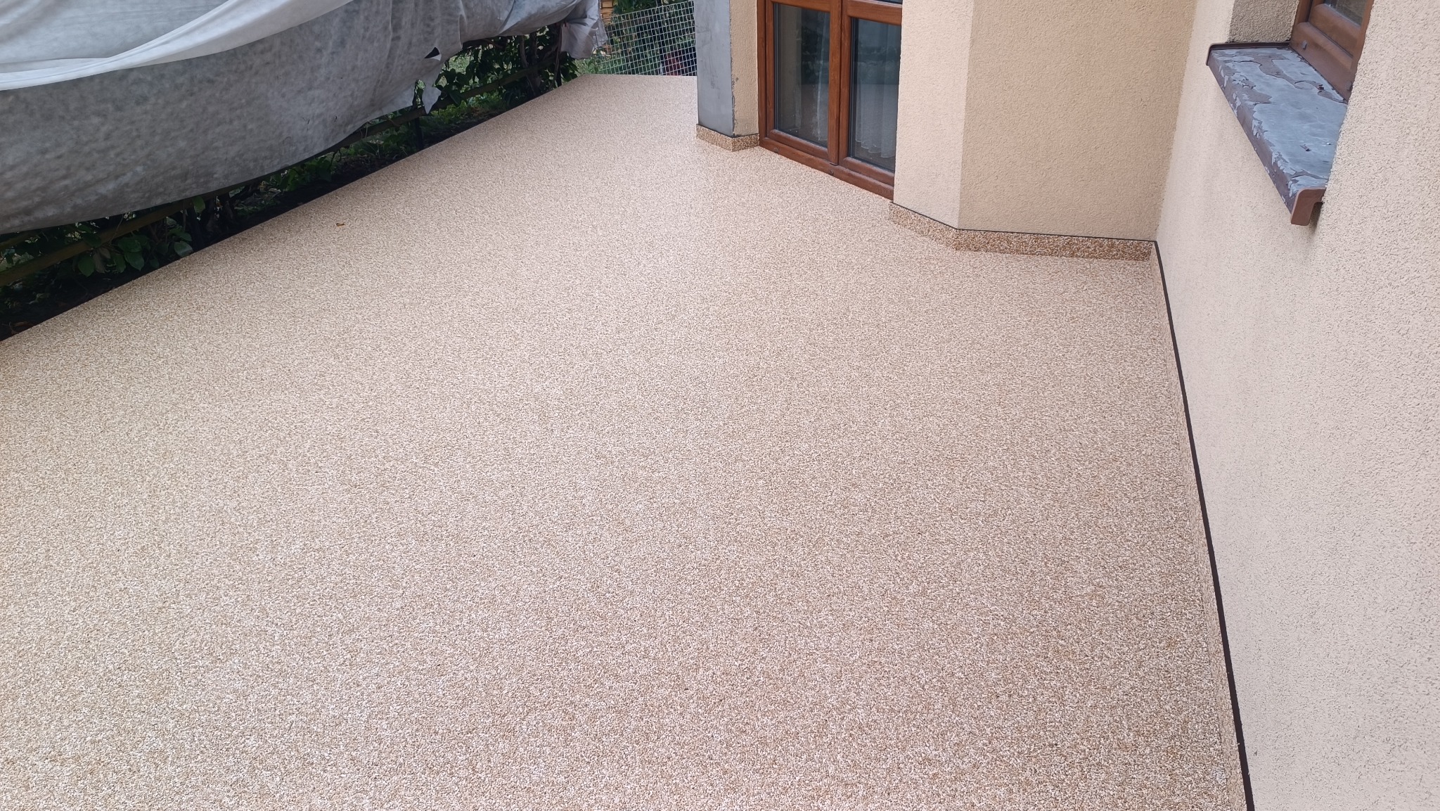 Beżowa, drobnoziarnista powierzchnia balkonu z wykończeniem przy ścianie budynku i oknach, częściowo zadaszona, widok z góry.