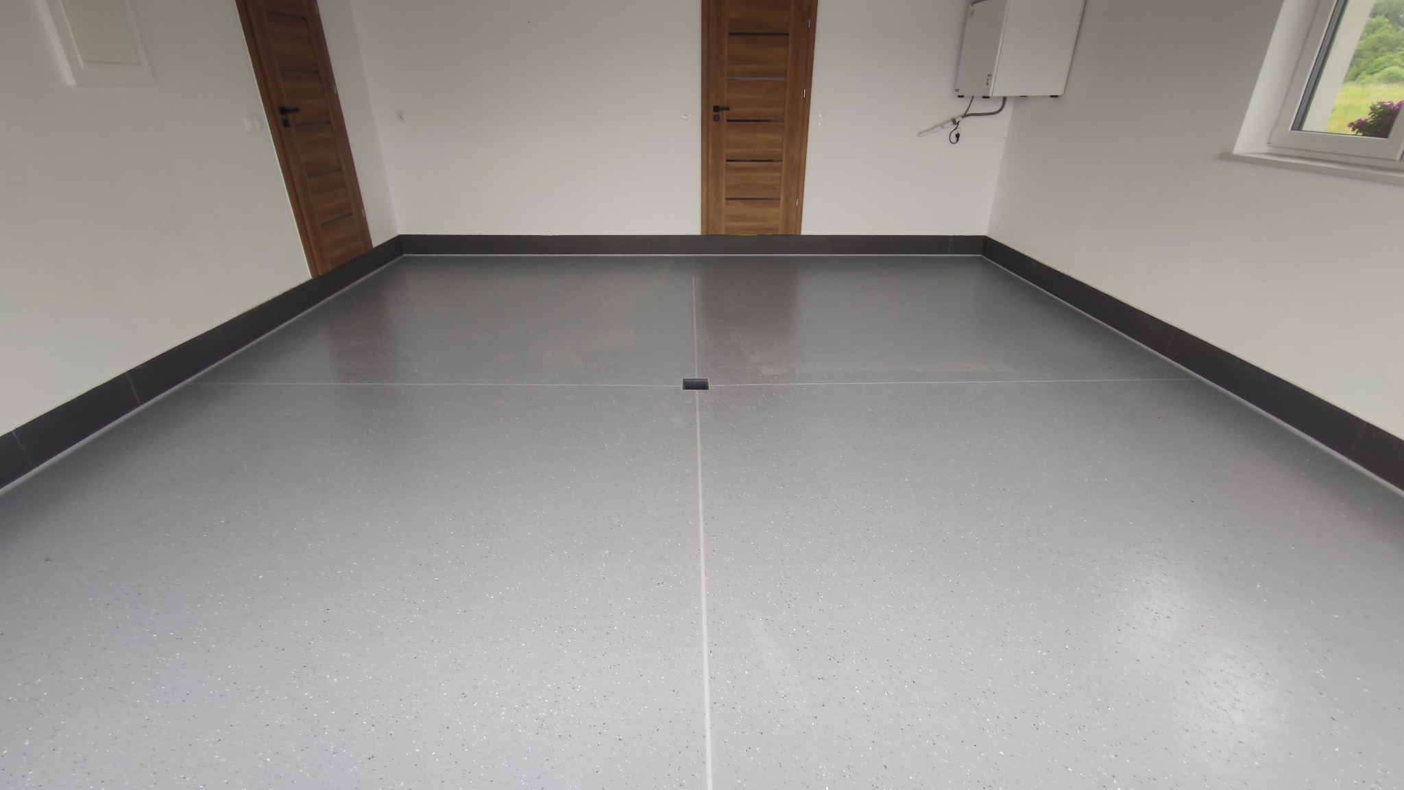 Szara posadzka z drobnymi plamkami w garażu, wykończona czarną listwą przypodłogową. Widoczne dwie drewniane drzwi i okno z widokiem na zieleń.