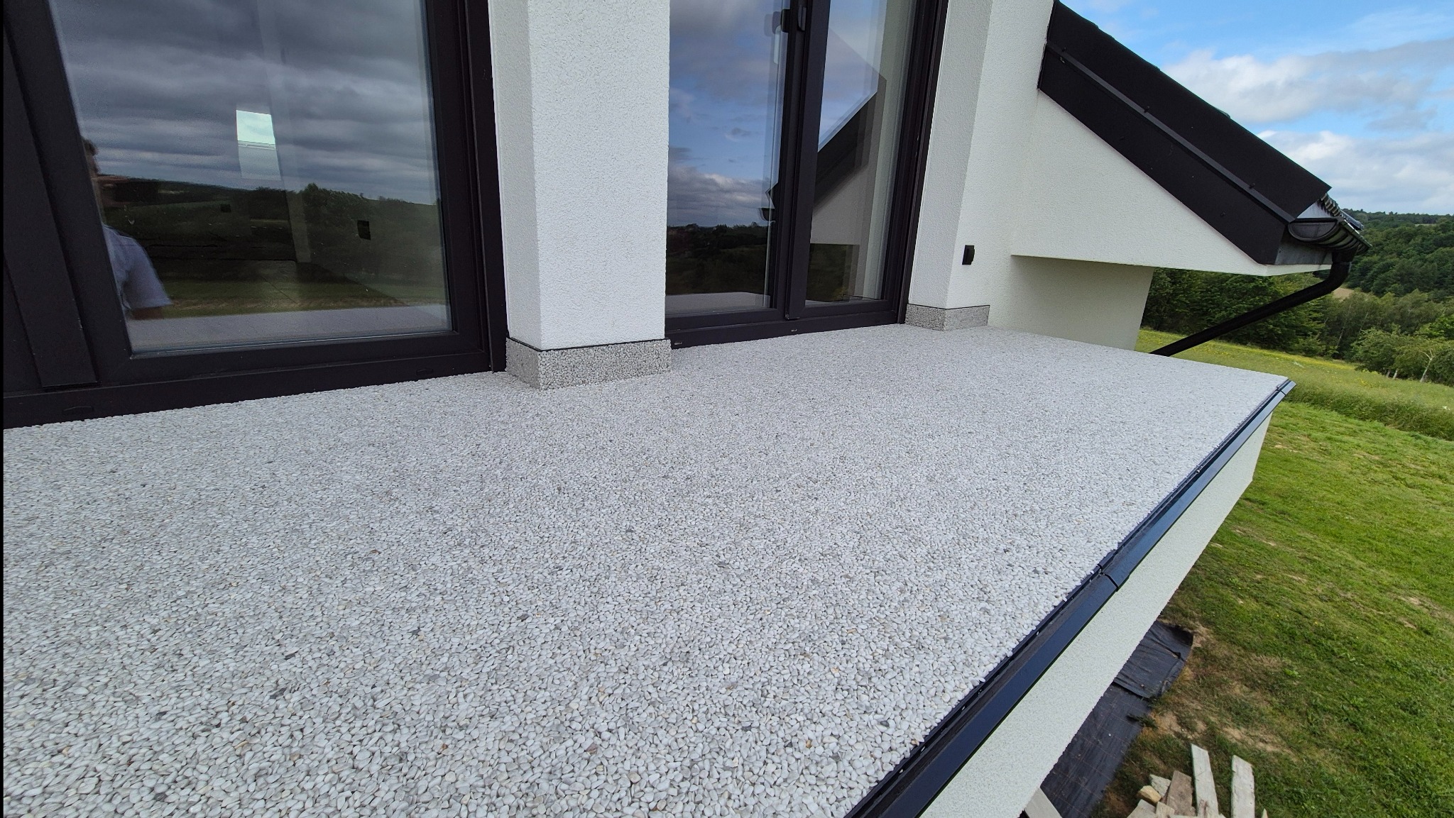 Jasnoszara posadzka z kamyczków na balkonie nowoczesnego domu z widokiem na zieleń. Minimalistyczny design, czarne ramy okienne, białe ściany.