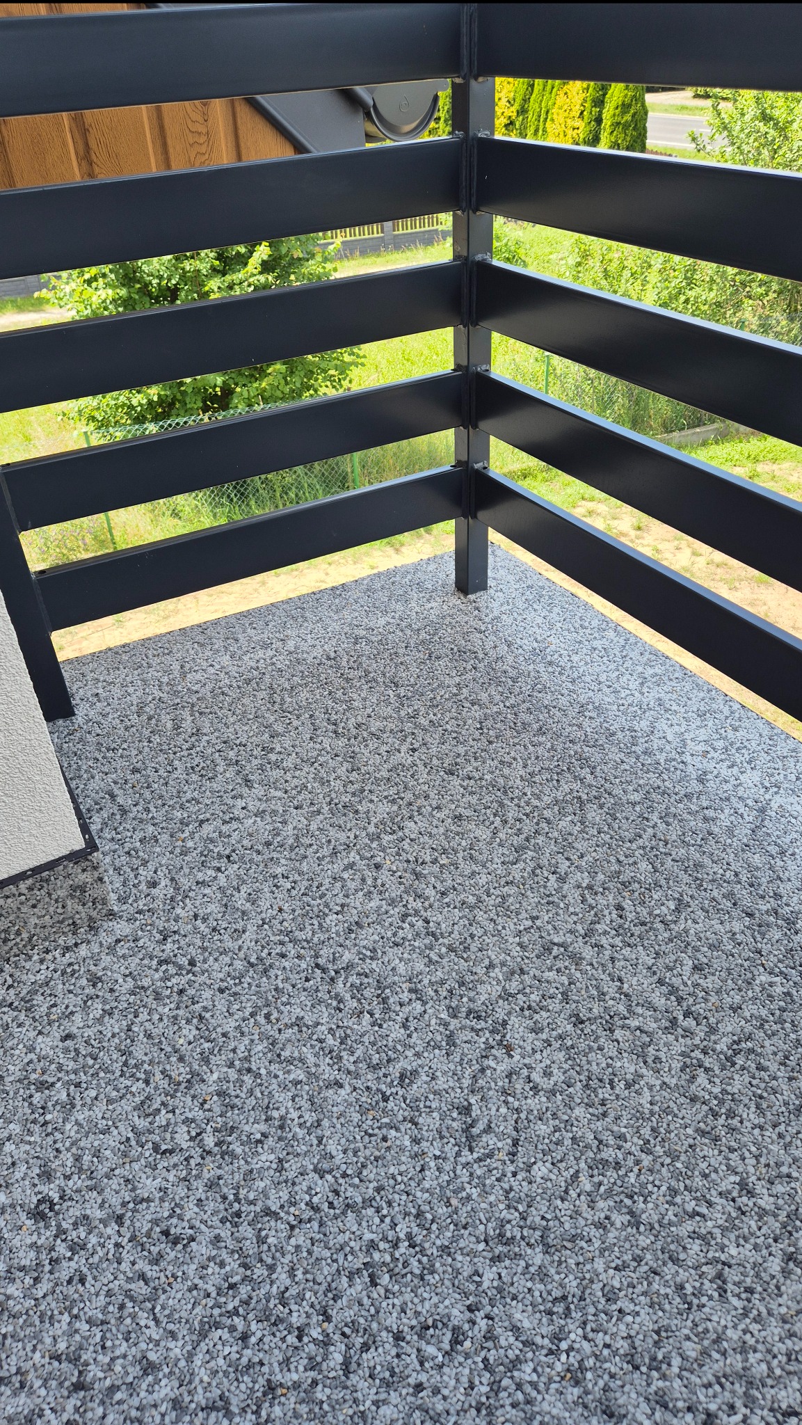 Szara posadzka z kamyczków na balkonie z czarną, metalową balustradą. Widok na zieleń za barierką. Nowoczesne wykończenie.