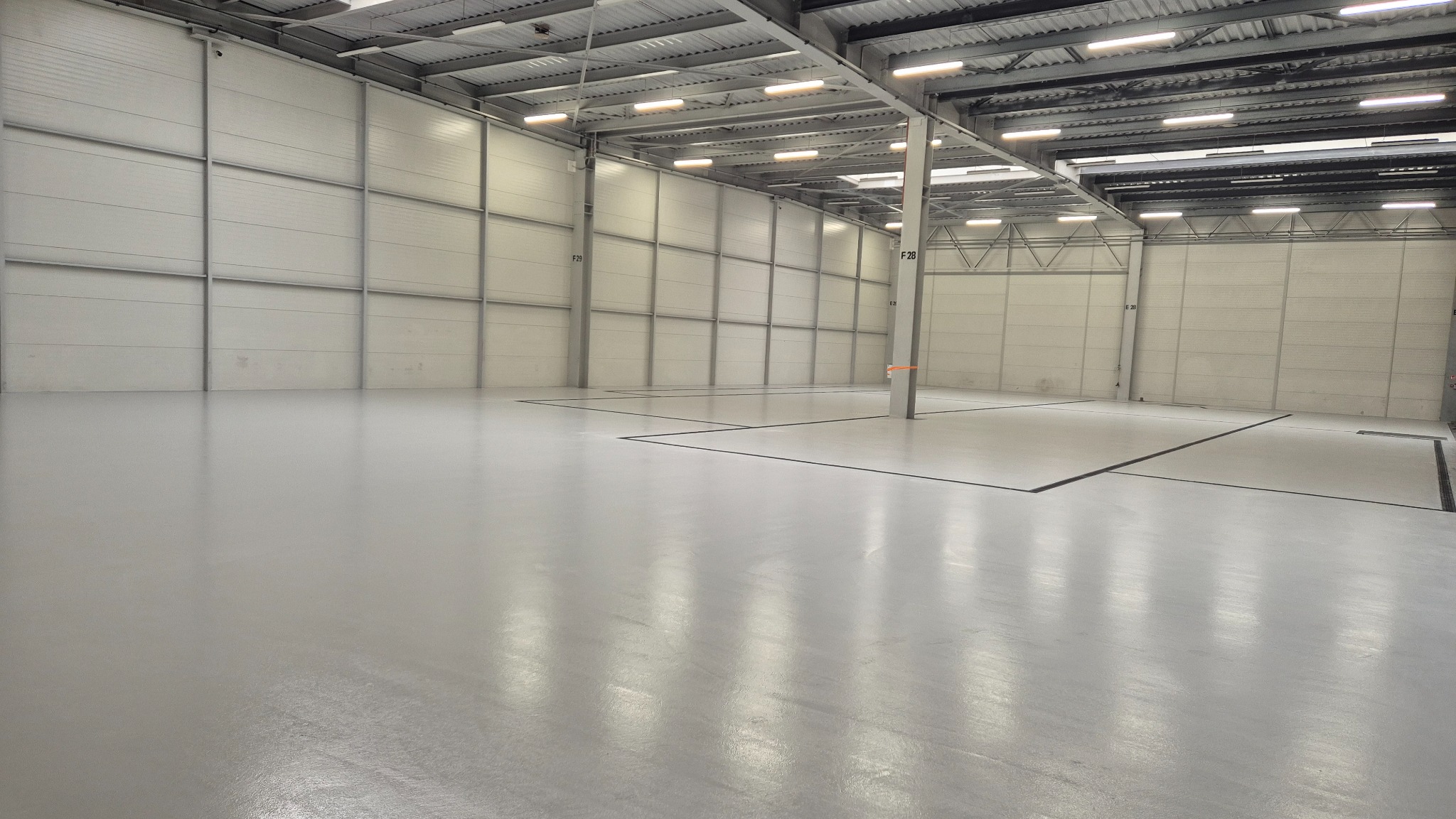 Jasnoszara, gładka posadzka w hali magazynowej z panelowymi ścianami i metalową konstrukcją dachu. Widoczne czarne linie podziału na posadzce i numery na ścianach.