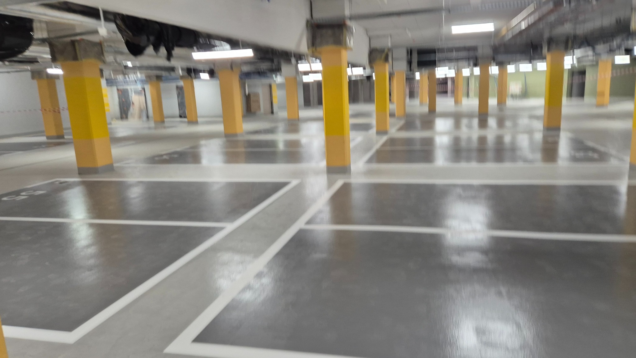 Błyszcząca, szara posadzka w garażu podziemnym z żółtymi słupami. Widoczne białe linie wyznaczające miejsca parkingowe. Oświetlenie sufitowe odbija się w gładkiej powierzchni.