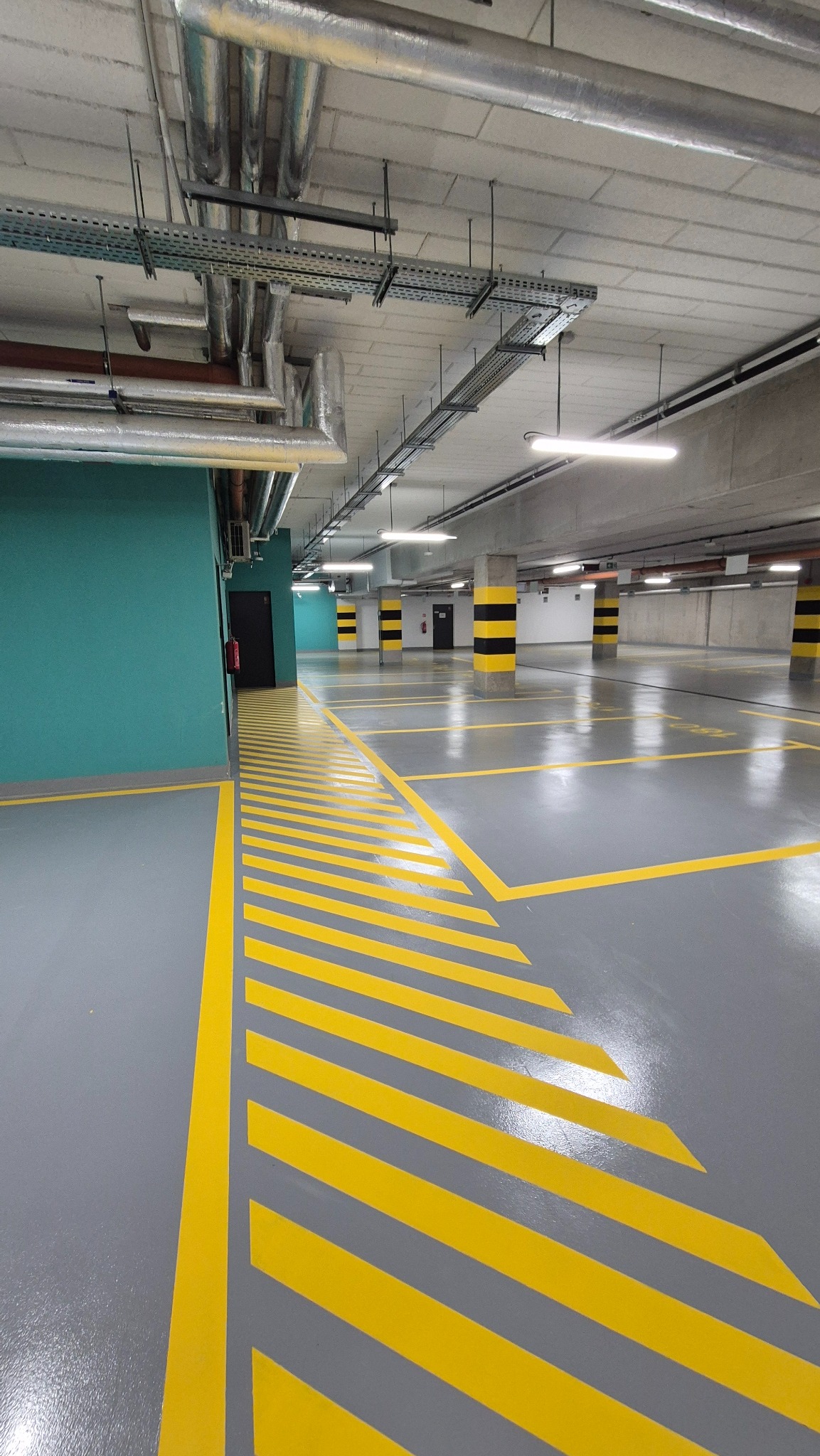 Szara posadzka w garażu podziemnym z żółtymi pasami i oznaczeniami. Widoczne rury pod sufitem i słupy w czarno-żółte pasy. Ściana w kolorze morskim.