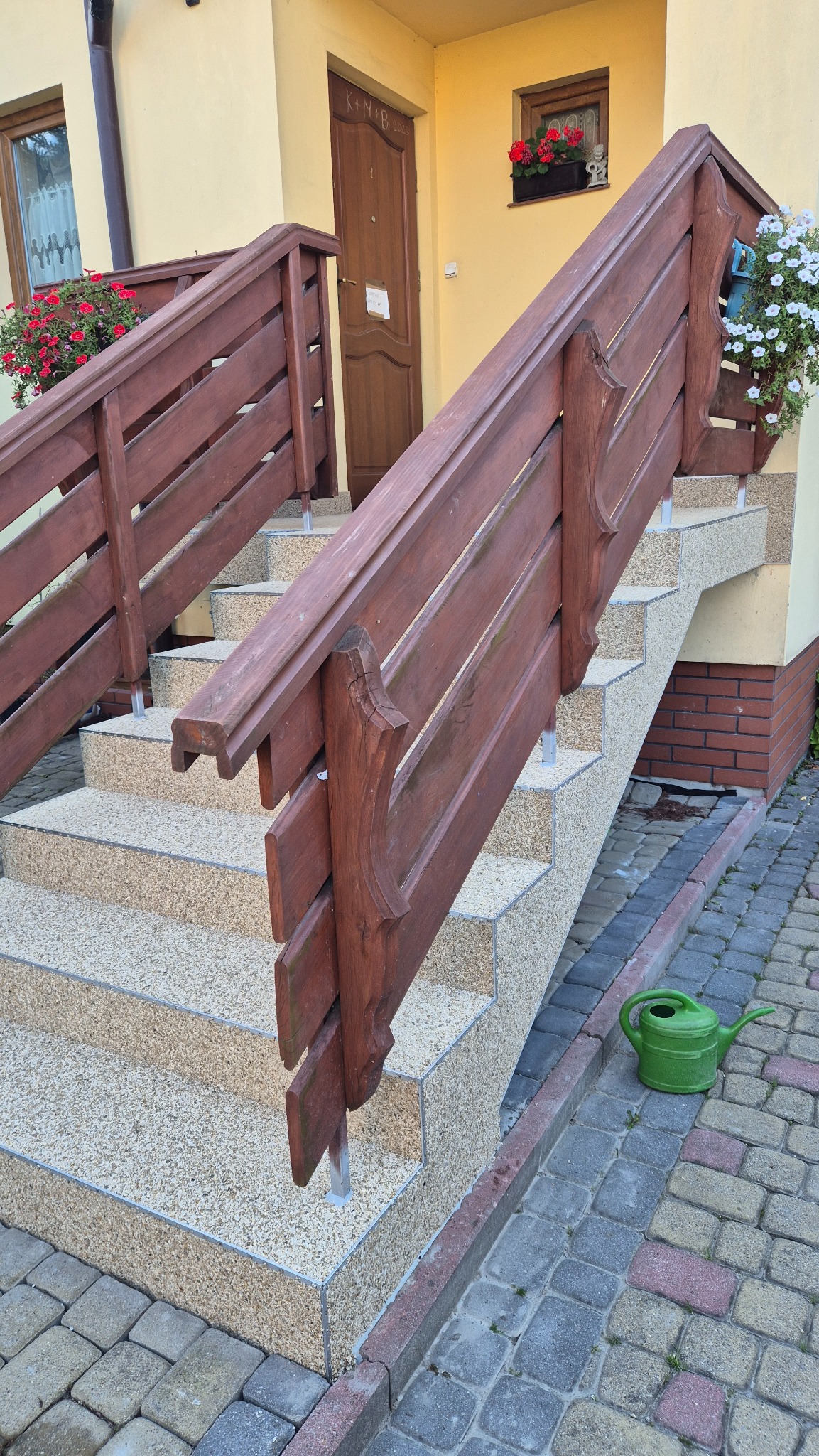 Schody wejściowe z drewnianą balustradą, wykończone jasnym, drobnym kamieniem. Na dole zielona konewka, w tle kwiaty w oknach i drzwi wejściowe.