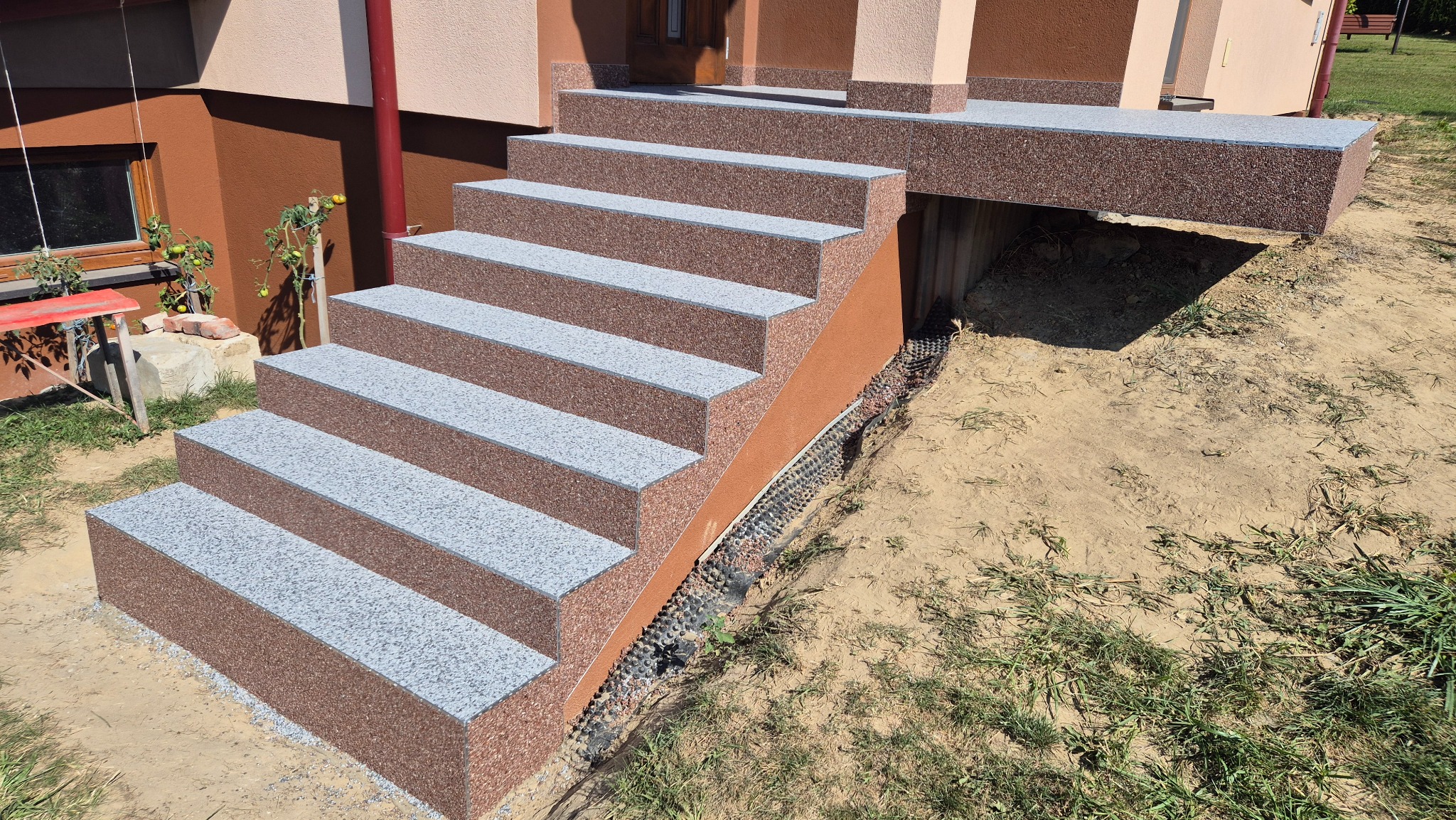 Schody wejściowe wykończone brązowo-szarą posadzką z żywicy, prowadzące do domu z brązową elewacją. Widoczna drenażowa warstwa przy schodach.