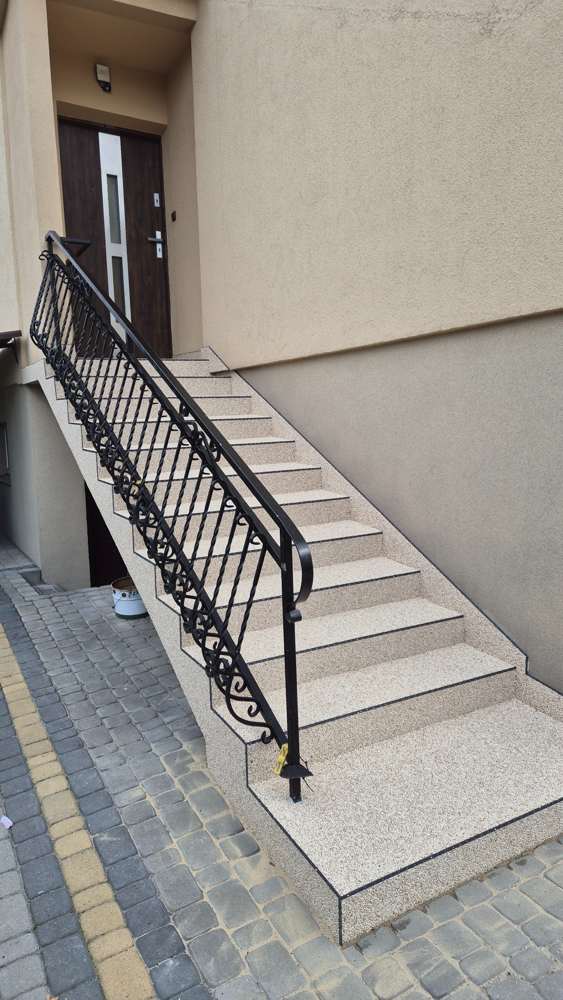 Zewnętrzne schody z beżową, kamyczkową nawierzchnią i czarną, kutą balustradą prowadzą do drzwi wejściowych. Widoczna kostka brukowa i fragment elewacji budynku.