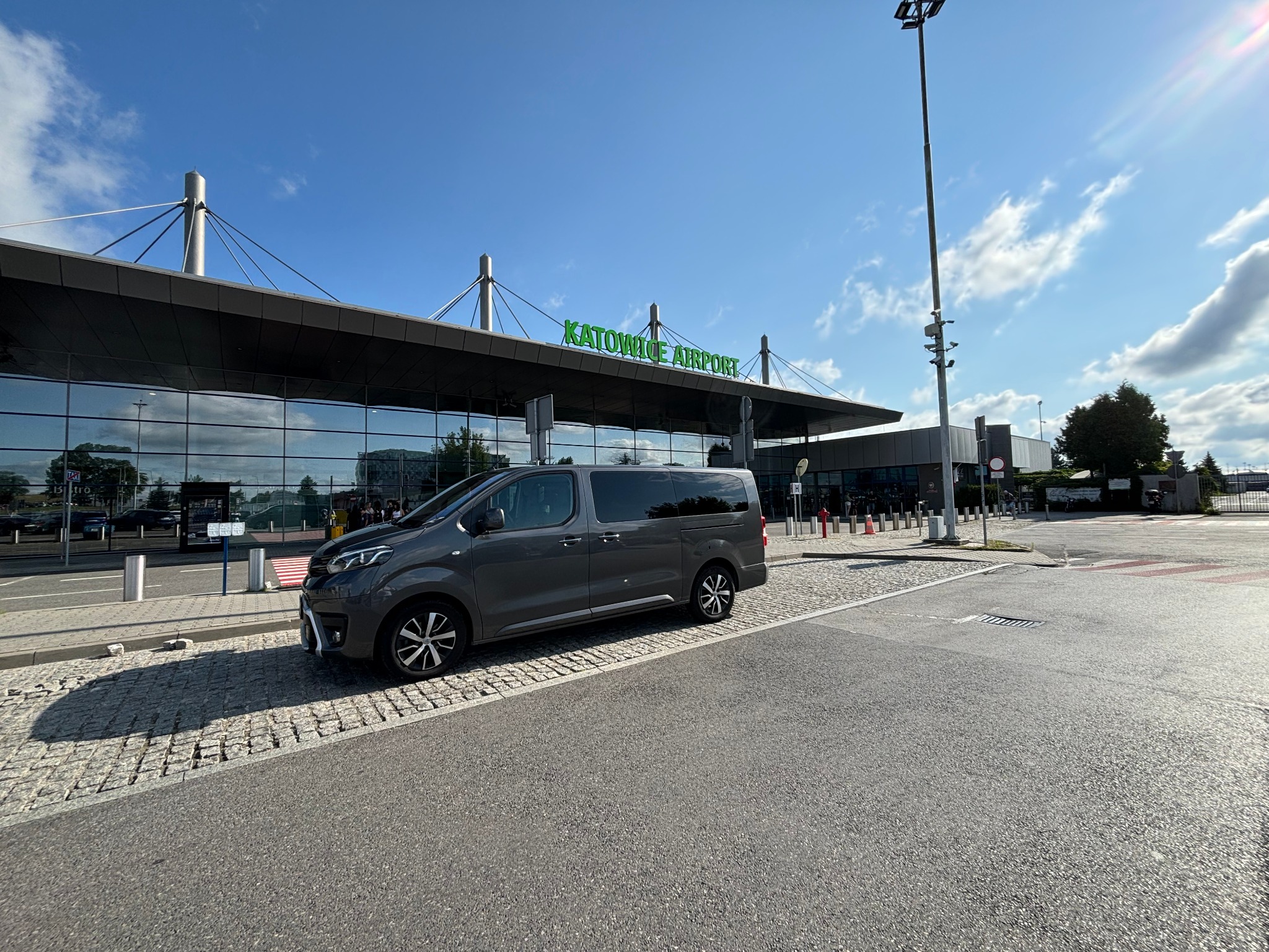 Szary bus marki Peugeot stoi zaparkowany przed budynkiem lotniska Katowice Airport w słoneczny dzień.