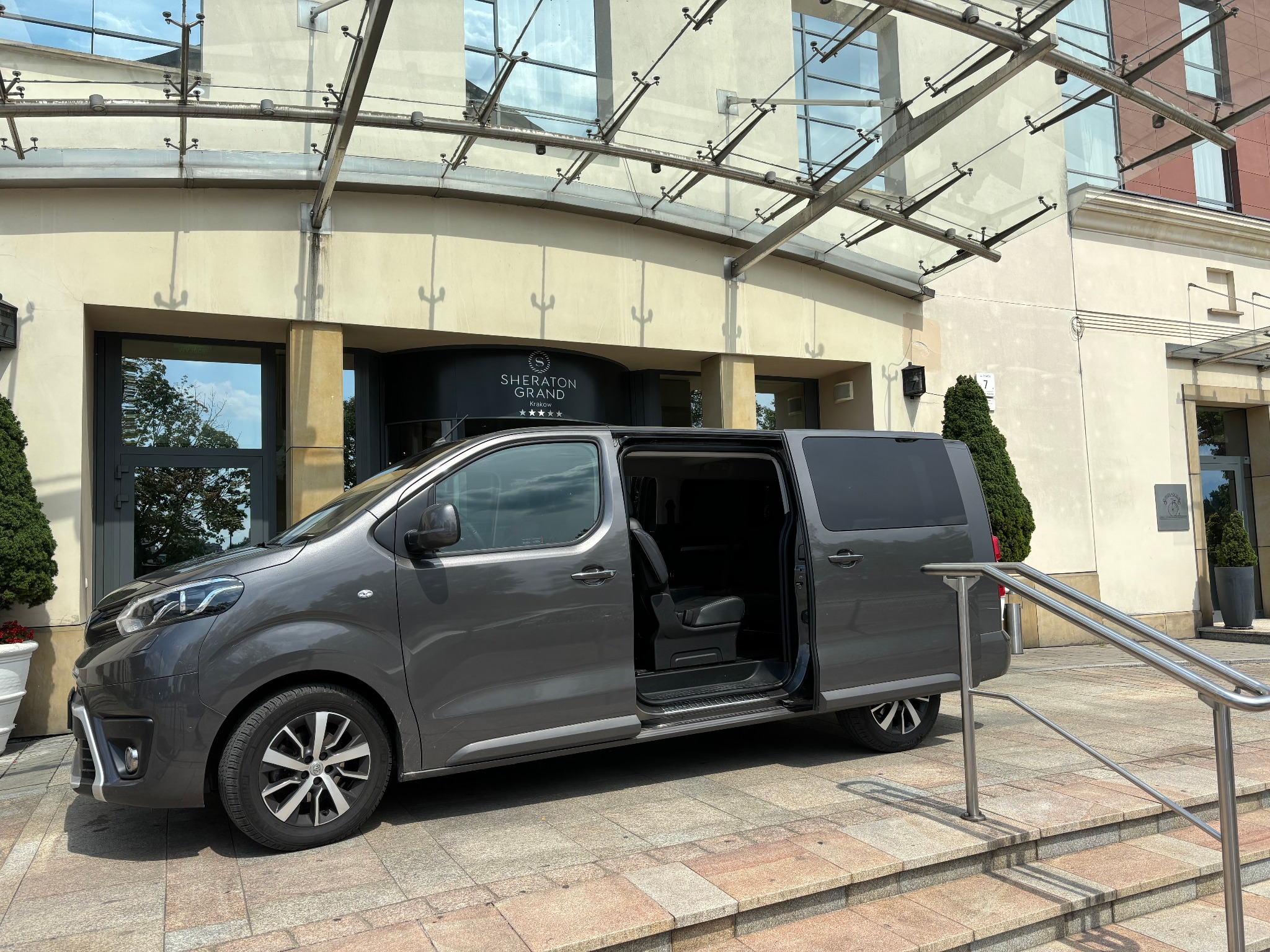Szary van z otwartymi drzwiami zaparkowany przed wejściem do hotelu Sheraton Grand w Krakowie, widoczny fragment schodów i balustrady.
