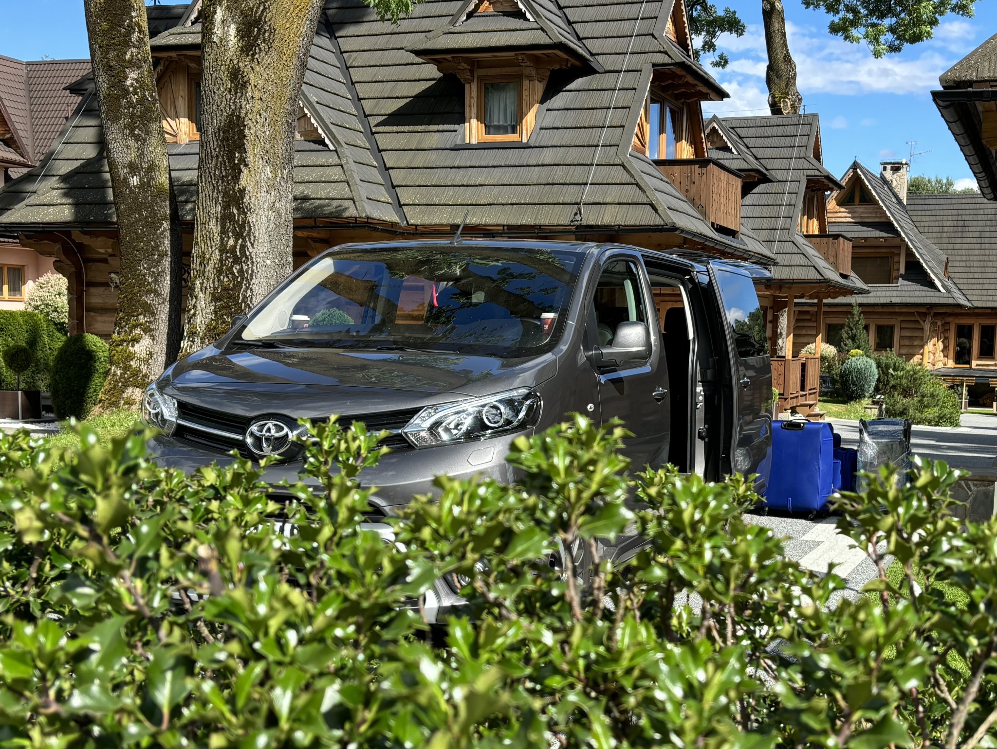 Szary van z otwartymi drzwiami na tle drewnianego domu w stylu góralskim, z widocznymi niebieskimi walizkami na zewnątrz, częściowo zasłonięty przez krzew.