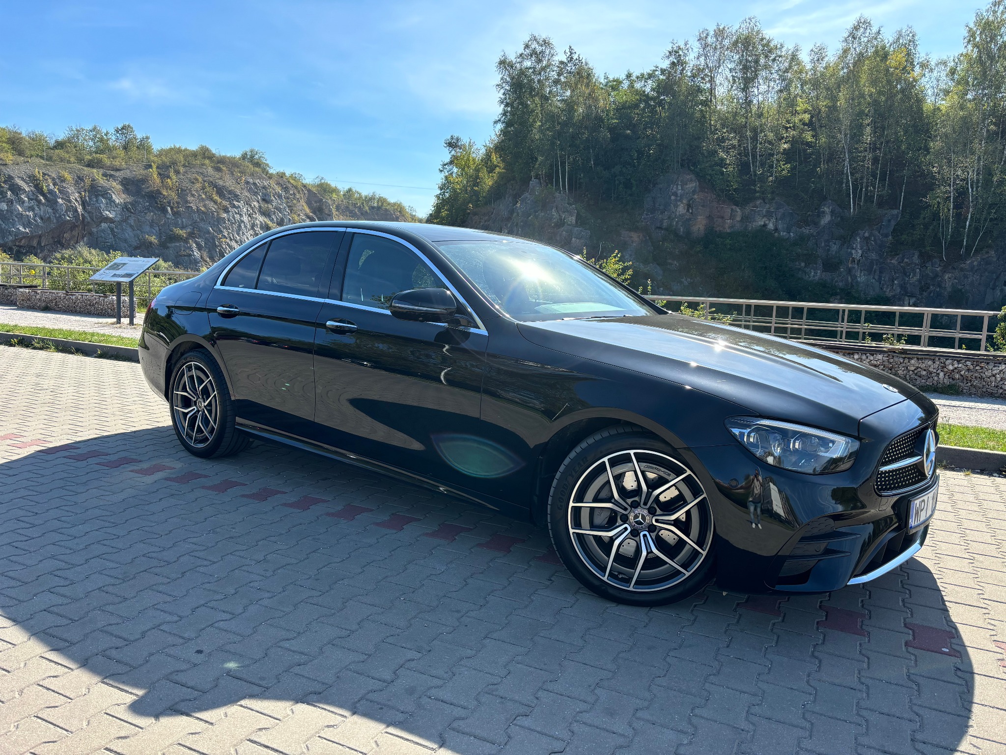 Czarny Mercedes-Benz klasy E zaparkowany na brukowanym placu, z widokiem na skaliste wzgórze i drzewa w tle, odbicie słońca na karoserii.