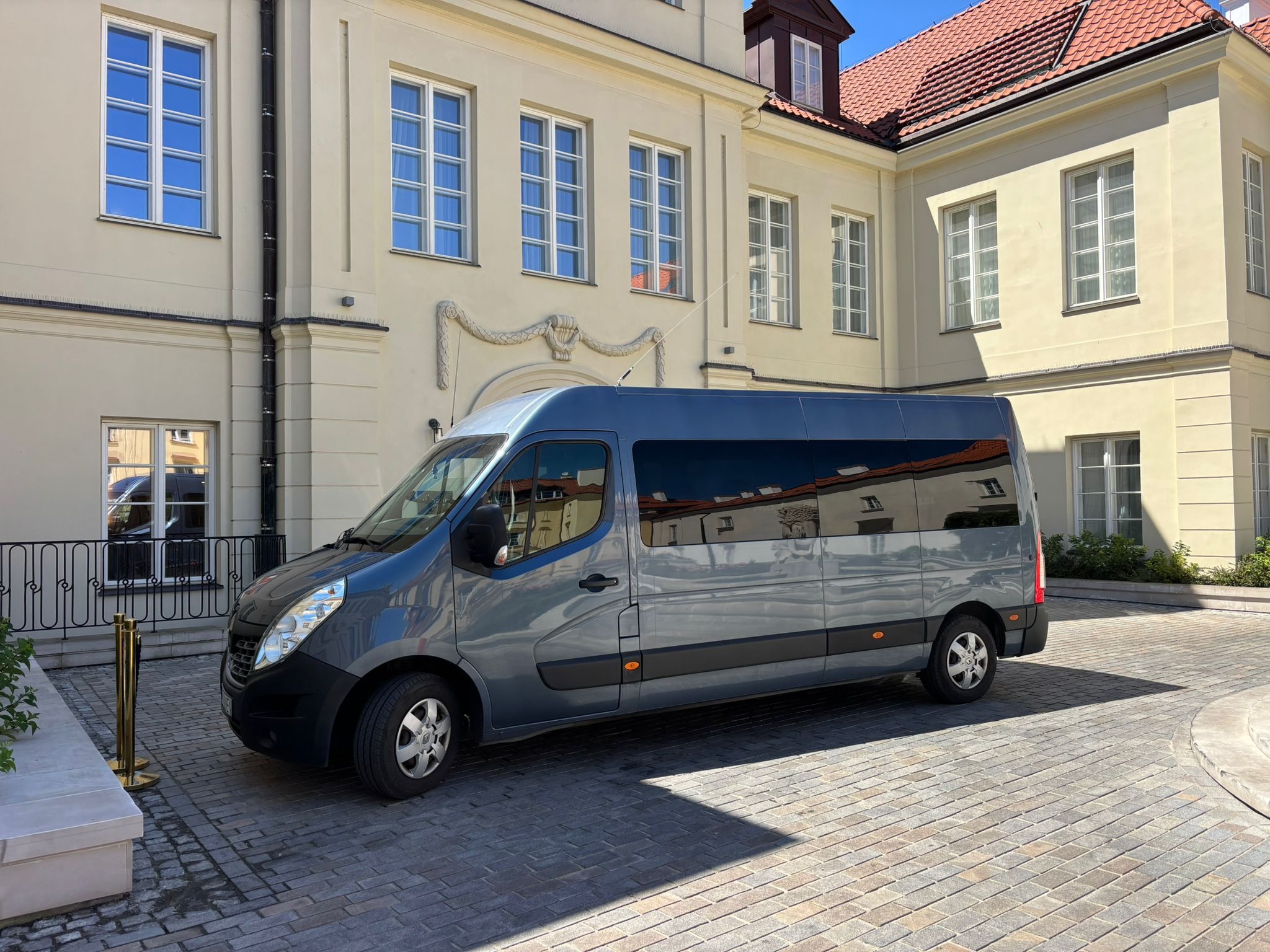 Szary bus Renault Master na tle klasycystycznego budynku z licznymi oknami. Pojazd zaparkowany na brukowanym dziedzińcu w słoneczny dzień.