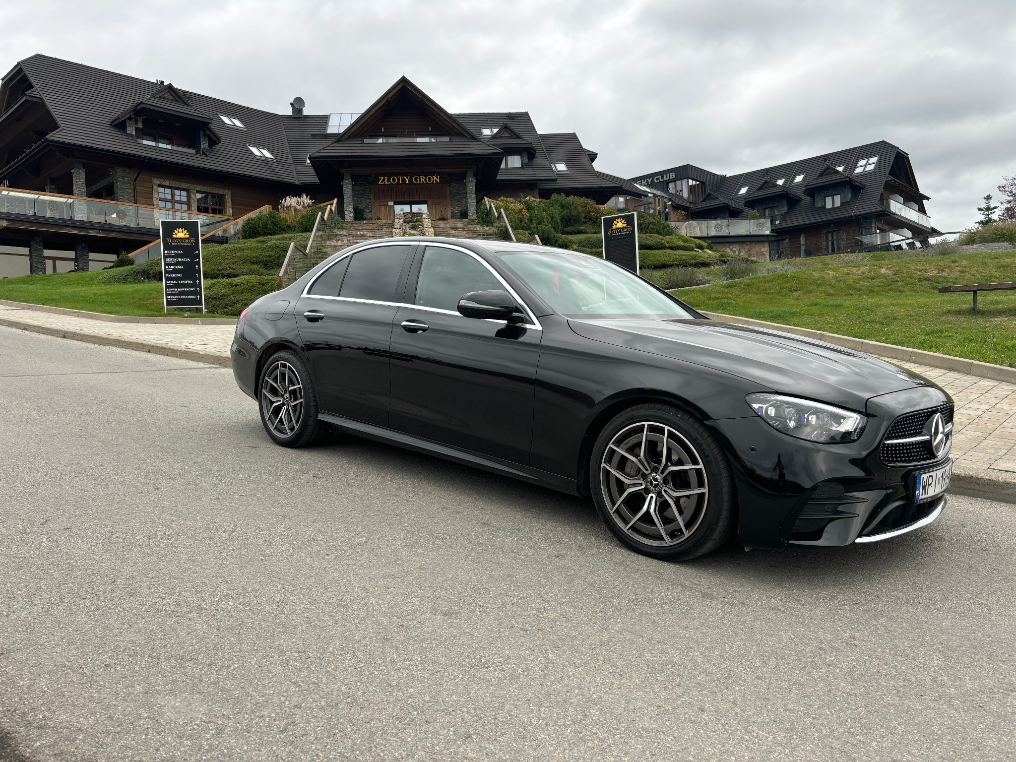 Czarna limuzyna Mercedes E-Klasa na tle górskiego hotelu Złoty Groń. Elegancki samochód przed luksusowym obiektem, pochmurne niebo.