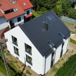 Nowoczesny dom z białą elewacją i grafitowym dachem z oknami dachowymi, widok z góry, w tle zabudowa jednorodzinna i szklarnia.