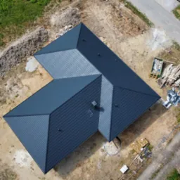 Widok z drona na nowo pokryty, grafitowy dach domu jednorodzinnego z kominem, na działce w trakcie zagospodarowywania; widoczne materiały budowlane i śmieci.