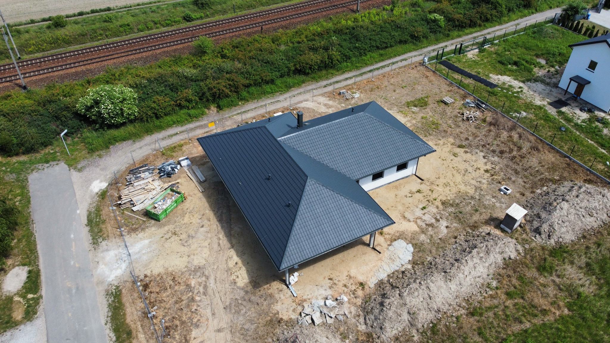 Widok z drona na dom jednorodzinny z ciemnoszarą, nową dachówką, w trakcie prac wykończeniowych na posesji, z widocznym kontenerem na odpady budowlane i torami kolejowymi w tle.