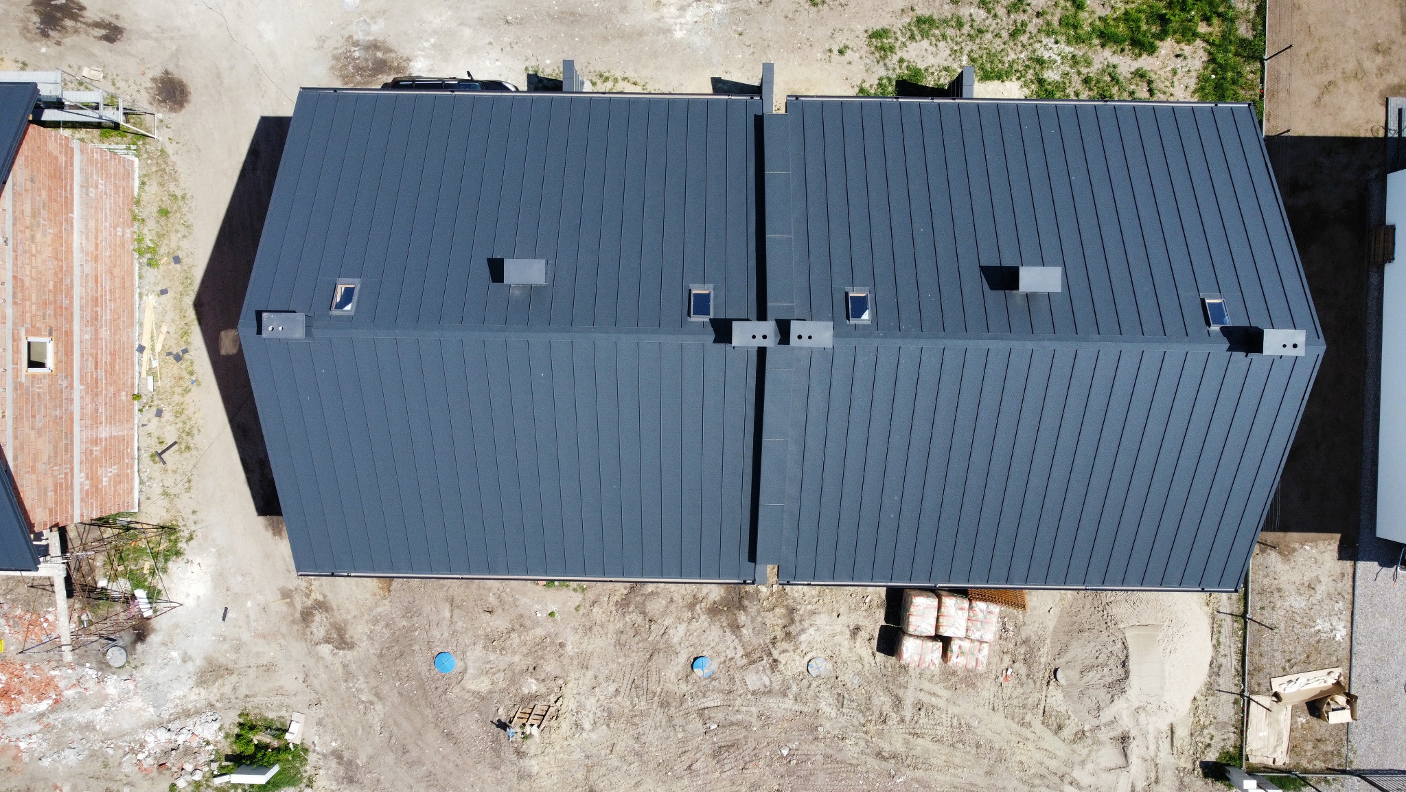 Widok z góry na ukończony, grafitowy dach dwuspadowy z blachy na bliźniaku, z oknami dachowymi i kominami, teren wokół w trakcie prac wykończeniowych.