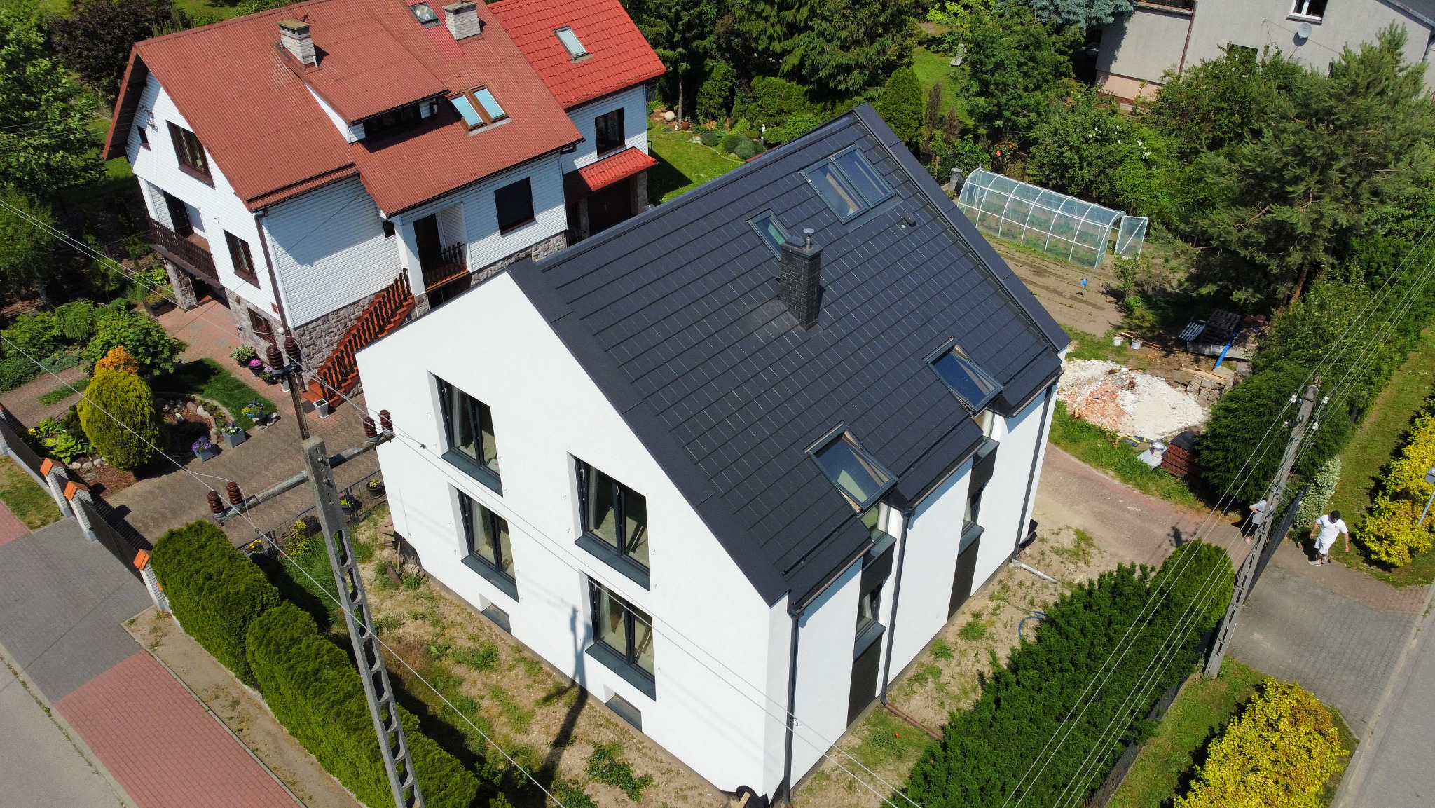Nowoczesny dom z białą elewacją i grafitowym dachem z oknami dachowymi, widok z góry, w tle zabudowa jednorodzinna i szklarnia.