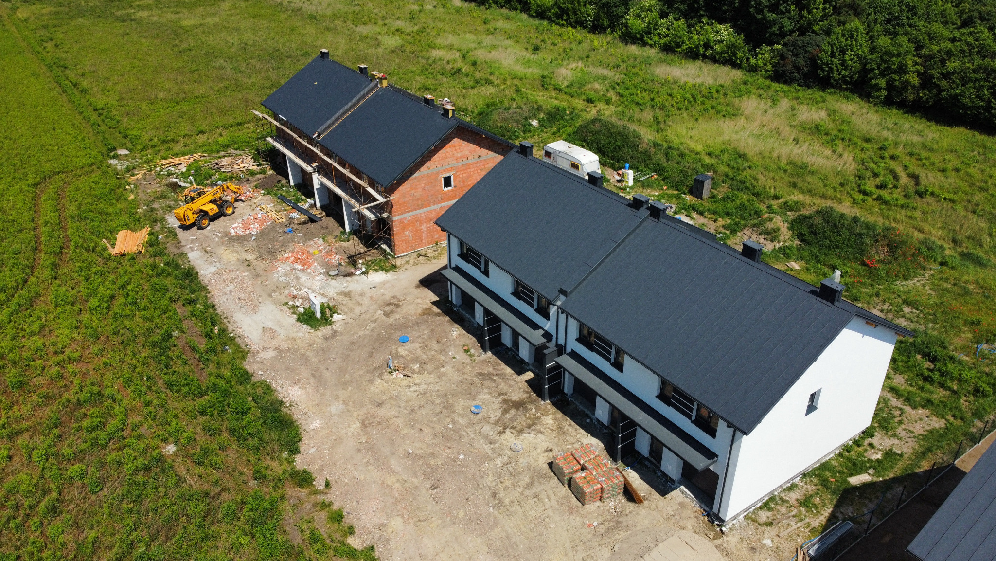 Widok z lotu ptaka na szereg domów w różnym stadium budowy, z nowymi, ciemnymi dachami, jeden dom w stanie surowym z cegły, na placu budowy żółta ładowarka teleskopowa i palety z cegłami.