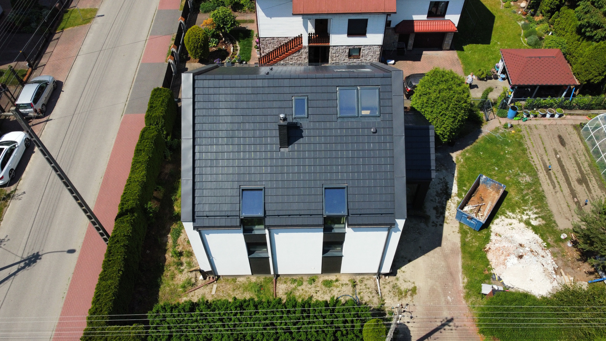 Widok z góry na dom z nowym, grafitowym dachem z dachówki ceramicznej z czterema oknami dachowymi i czarnym kominem, otoczony zielenią i zabudową jednorodzinną.