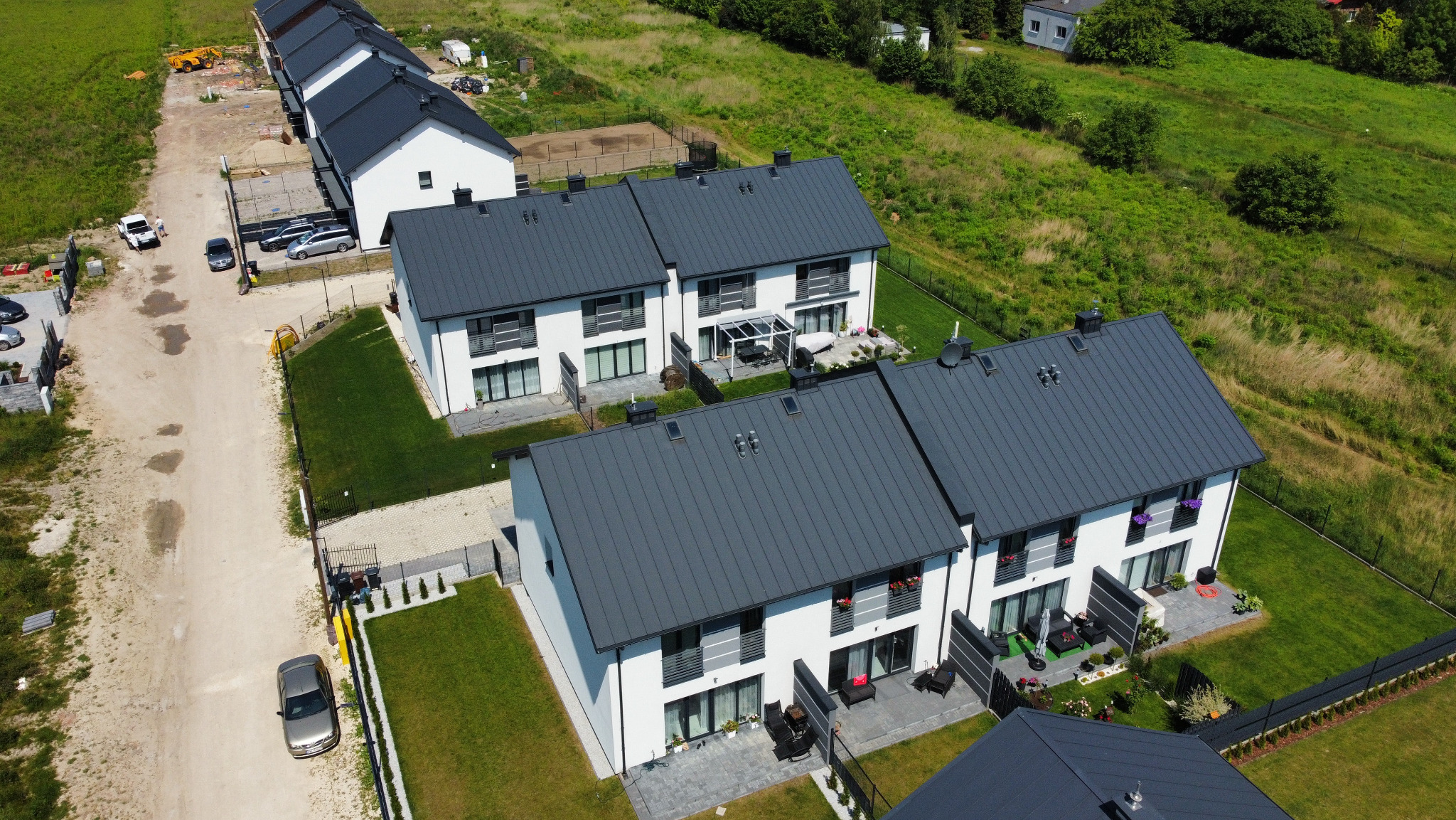 Widok z lotu ptaka na szereg nowych, dwukondygnacyjnych domów z szarymi dachami i białymi elewacjami, otoczonych zielonymi trawnikami i czarnymi ogrodzeniami, z widoczną drogą dojazdową w tle.