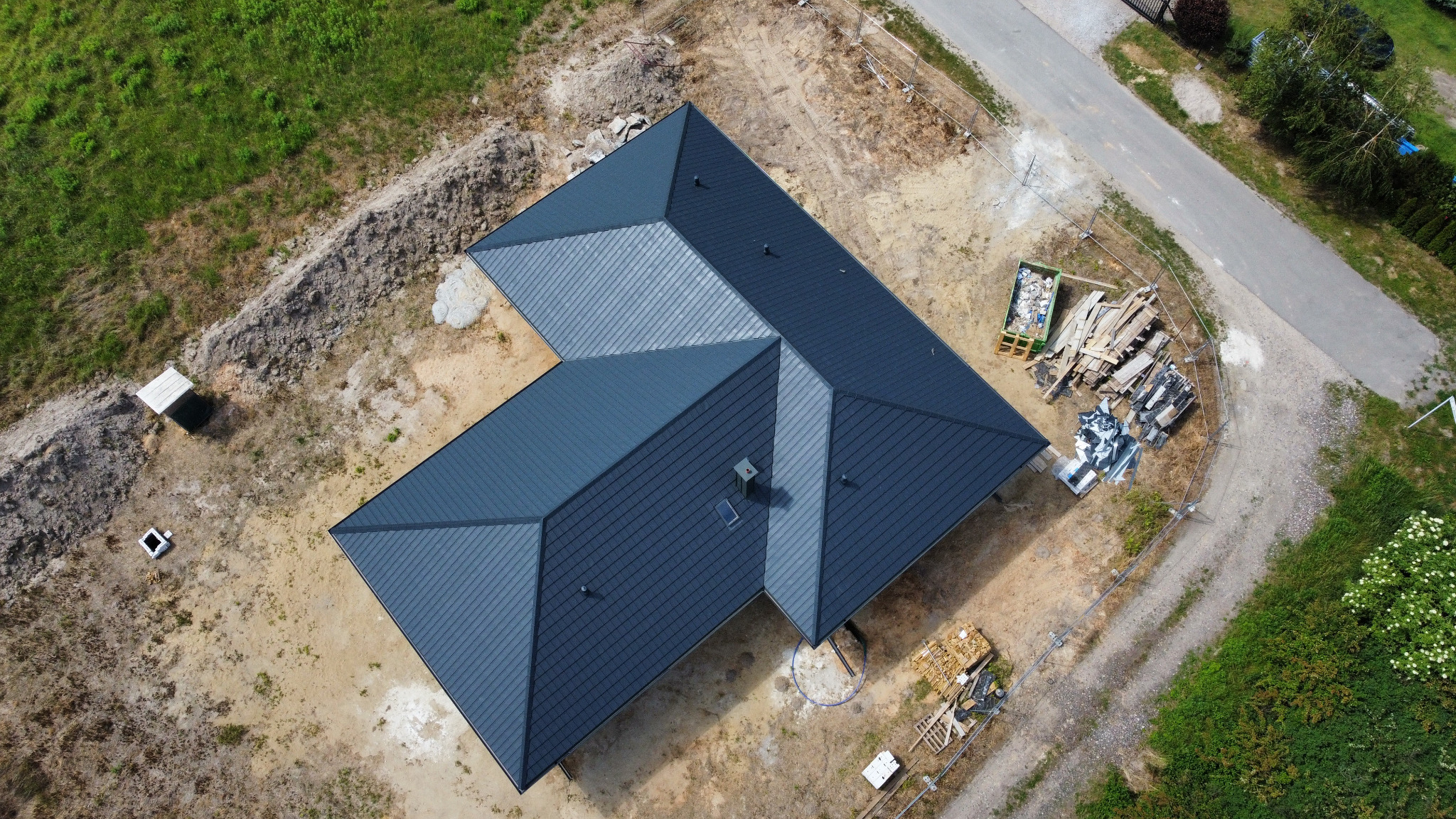 Widok z drona na nowo pokryty, grafitowy dach domu jednorodzinnego z kominem, na działce w trakcie zagospodarowywania; widoczne materiały budowlane i śmieci.