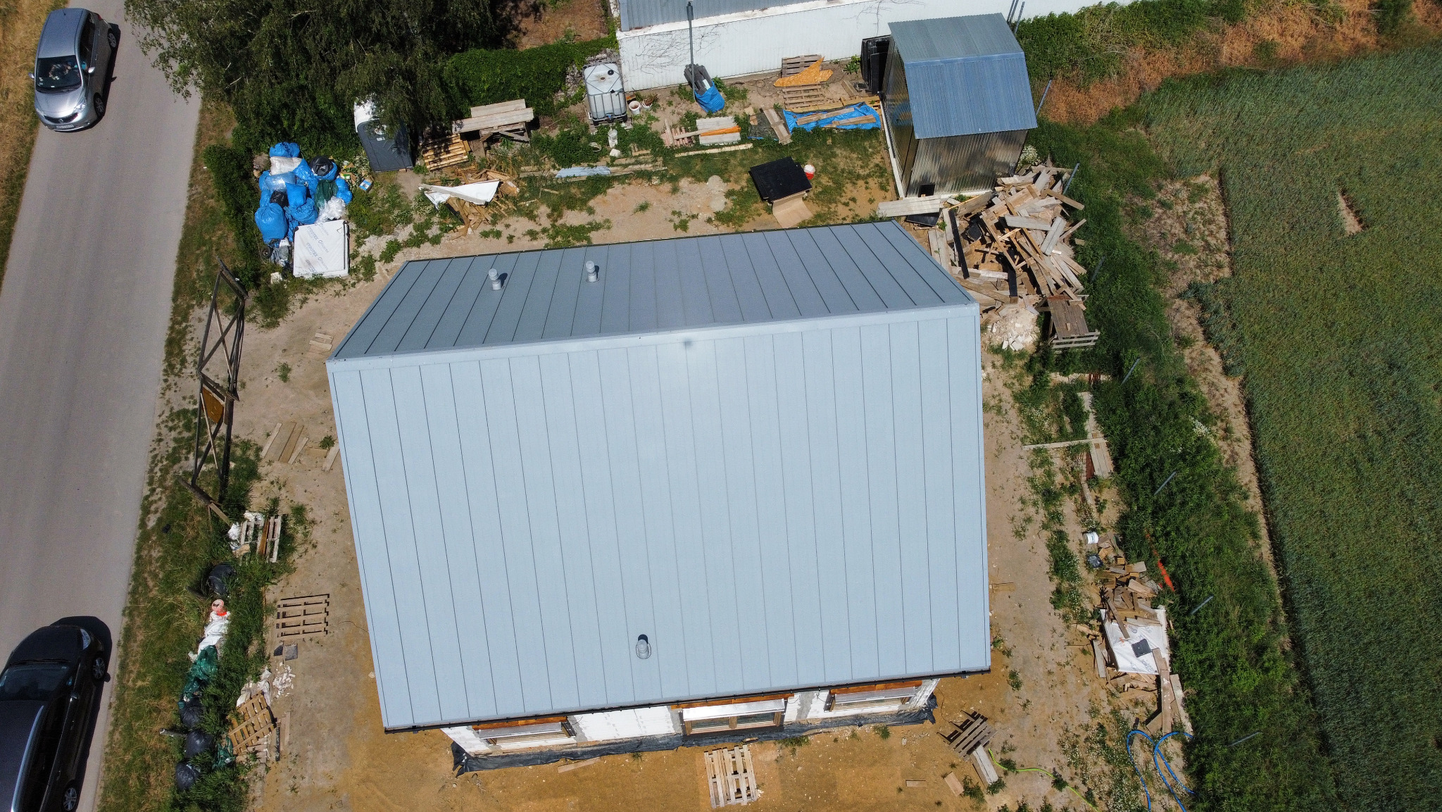 Widok z góry na budynek w trakcie budowy z nowym, metalowym dachem w kolorze jasnoszarym, na zewnątrz porozrzucane materiały budowlane, obok droga z zaparkowanymi samochodami, a w tle pole...