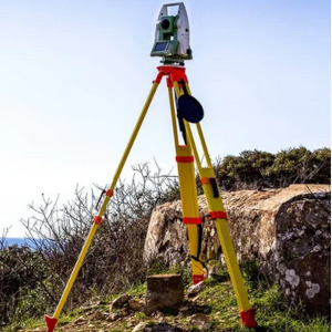 Zielono-szary tachimetr elektroniczny ustawiony na żółtym statywie z czerwonymi elementami mocującymi, na tle skalistego terenu i błękitnego nieba.