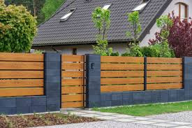 Nowoczesne ogrodzenie z poziomych desek w kolorze jasnego drewna, osadzone na ciemnych, grafitowych słupkach z bloczków betonowych, z furtką w tym samym stylu, na tle domu z szarym dachem i zieleni...