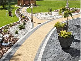 Zakrzywiona ścieżka z kostki brukowej w ogrodzie, z żółto-szarą nawierzchnią i obrzeżami z ciemnej kostki, prowadząca obok rabat z kamieniami i roślinami, oświetlona latarniami, z altaną w tle.