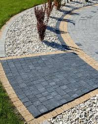 Kwadratowa powierzchnia wyłożona ciemnoszarą kostką brukową, otoczona obwódką z jasnej kostki, granicząca z białym żwirem i fragmentem ścieżki z szarej kostki. Całość wkomponowana w zielony trawnik...