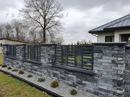 Nowoczesne ogrodzenie z szarej, łupanej imitacji kamienia z poziomymi, ciemnymi przęsłami drewnianymi, oddzielone niskim murkiem od trawnika z nasadzeniami kulistych krzewów i obrzeżem z kostki.