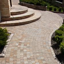 Kaskadowe schody z jasnej kostki brukowej prowadzące do domu, płynnie przechodzące w wybrukowaną ścieżkę, obrzeża wykończone ciemniejszym kamieniem, zieleń po bokach.