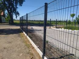 Czarne ogrodzenie z metalowej siatki panelowej na betonowej podmurówce, widoczne słupki i łączenia paneli, w tle trawnik i drzewa.