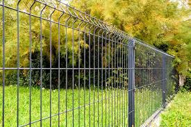 Czarne, metalowe ogrodzenie panelowe na tle soczystej zieleni trawnika i drzew. Widoczne pionowe pręty i poziome wzmocnienia. Perspektywa z dołu, uwydatniająca wysokość ogrodzenia.