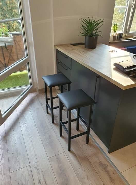 Nowoczesna wyspa kuchenna z blatem imitującym drewno, czarnymi szafkami i dwoma hokerami na tle jasnej podłogi i okna z widokiem na zieleń.